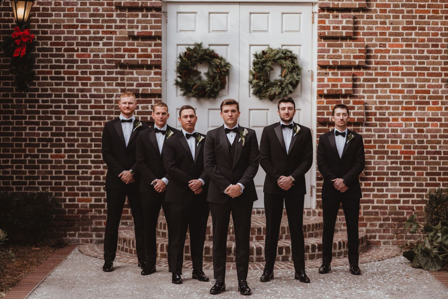 Groom and five groomsmen in matching black tuxedos with bow ties posing before brick wall with wreaths.