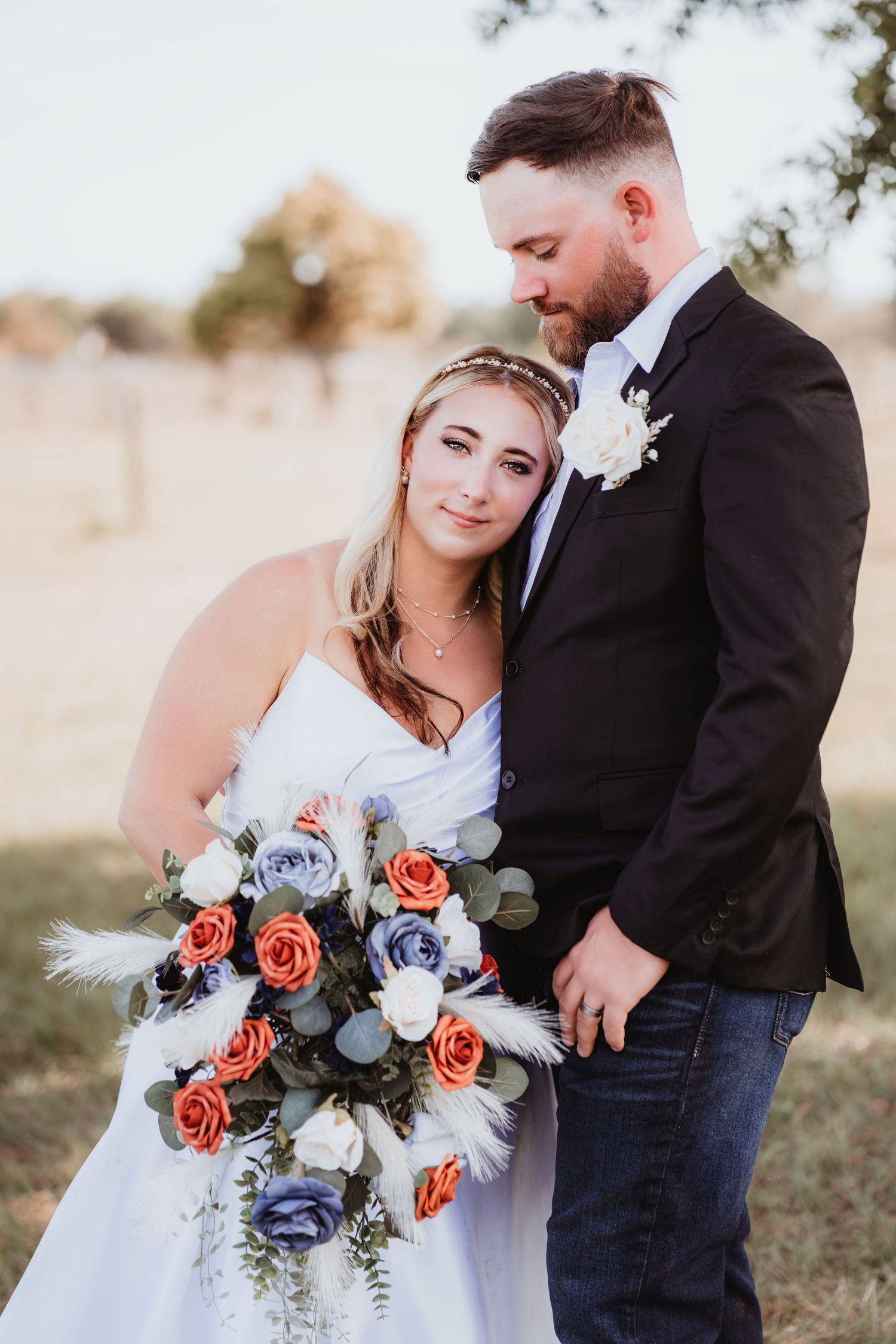 Groom in black suit gazing down at bride holding orange and blue floral bouquet in rustic setting.