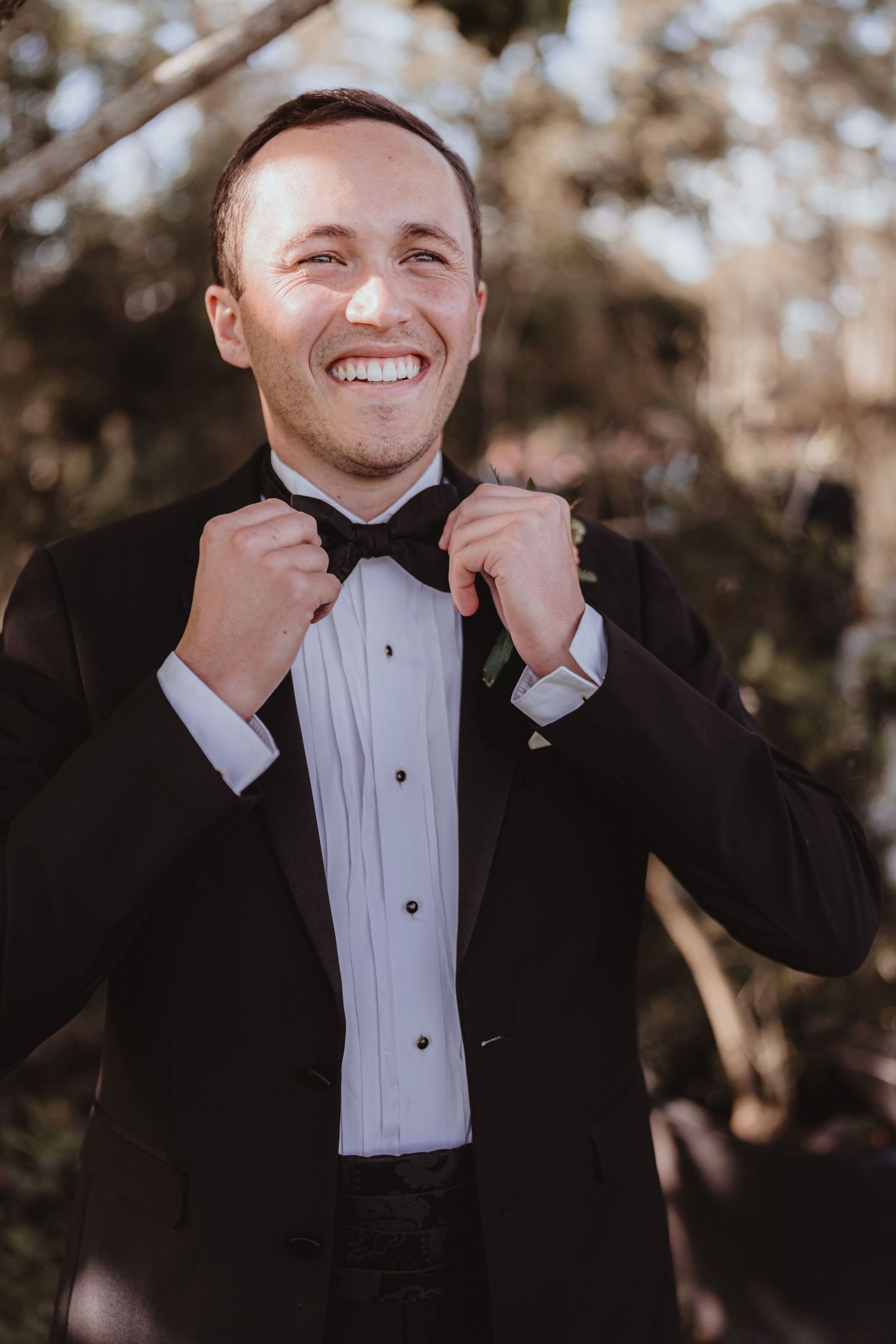 Smiling groom in black tuxedo adjusting his bow tie outdoors on his wedding day.