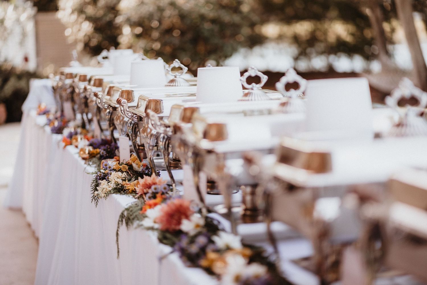 Outdoor wedding buffet table decorated with colorful flowers and chafing dishes.