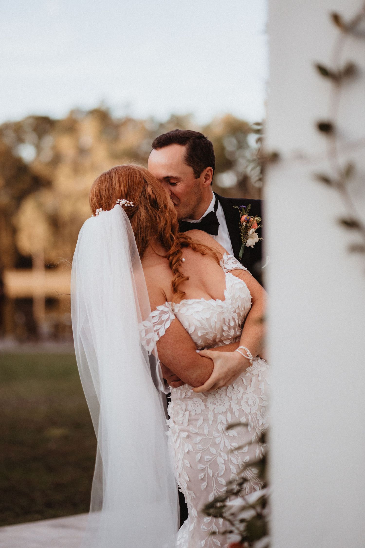 Groom in black tuxedo kisses bride in off-shoulder lace gown beside a white column at golden hour.