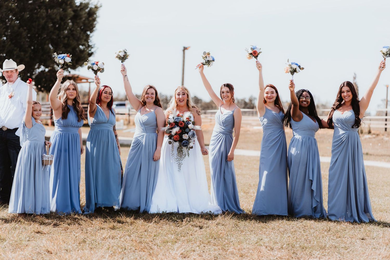 Bride in white gown with seven bridesmaids in blue dresses celebrating outdoors at a rustic wedding venue.