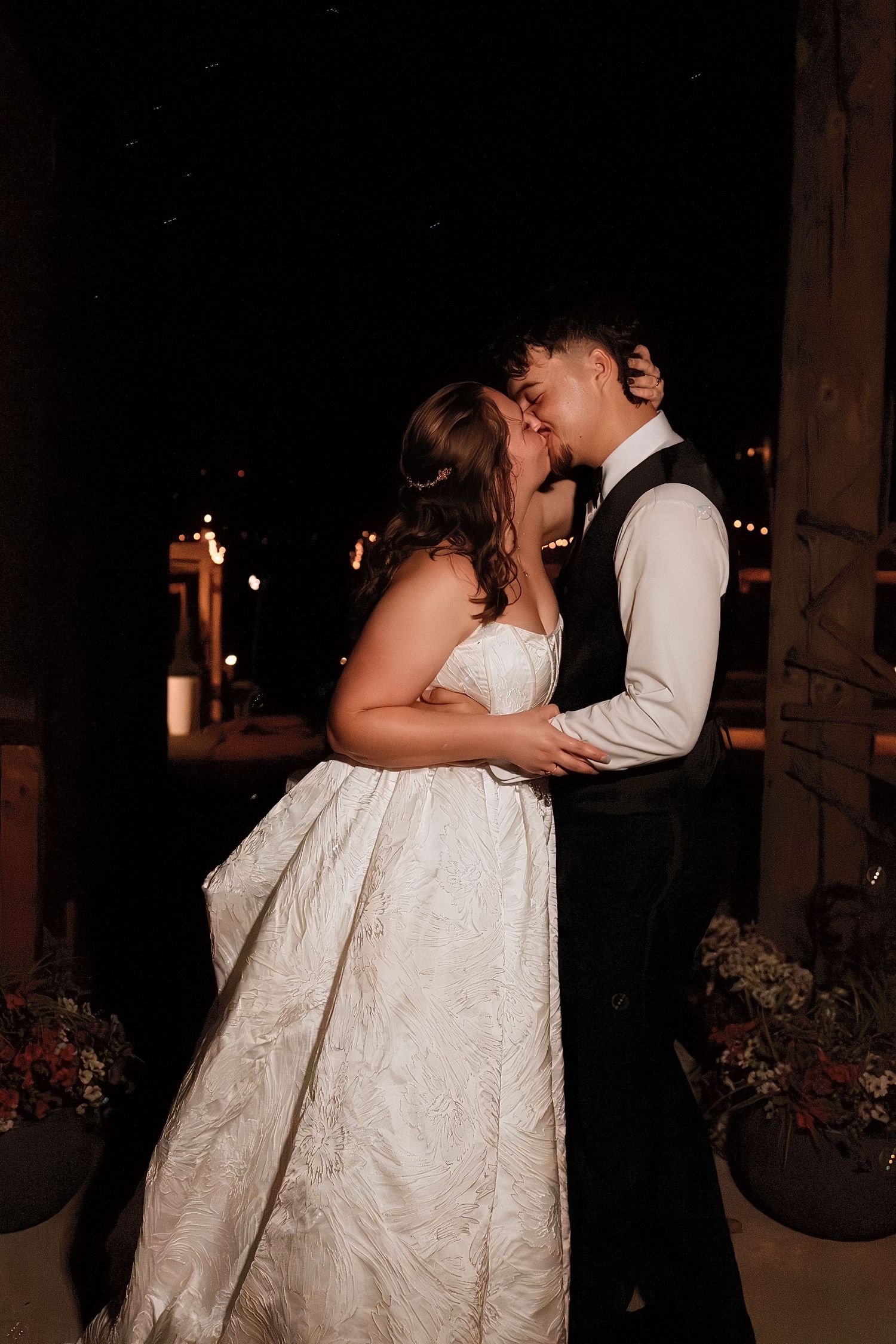 Bride and groom share a romantic kiss under string lights at dark outdoor wedding venue at night.