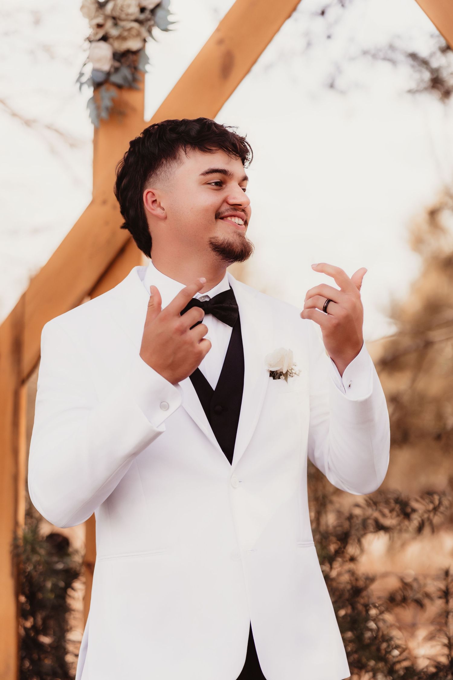Confident groom in white tuxedo with black bow tie posing near wooden arch at outdoor wedding.