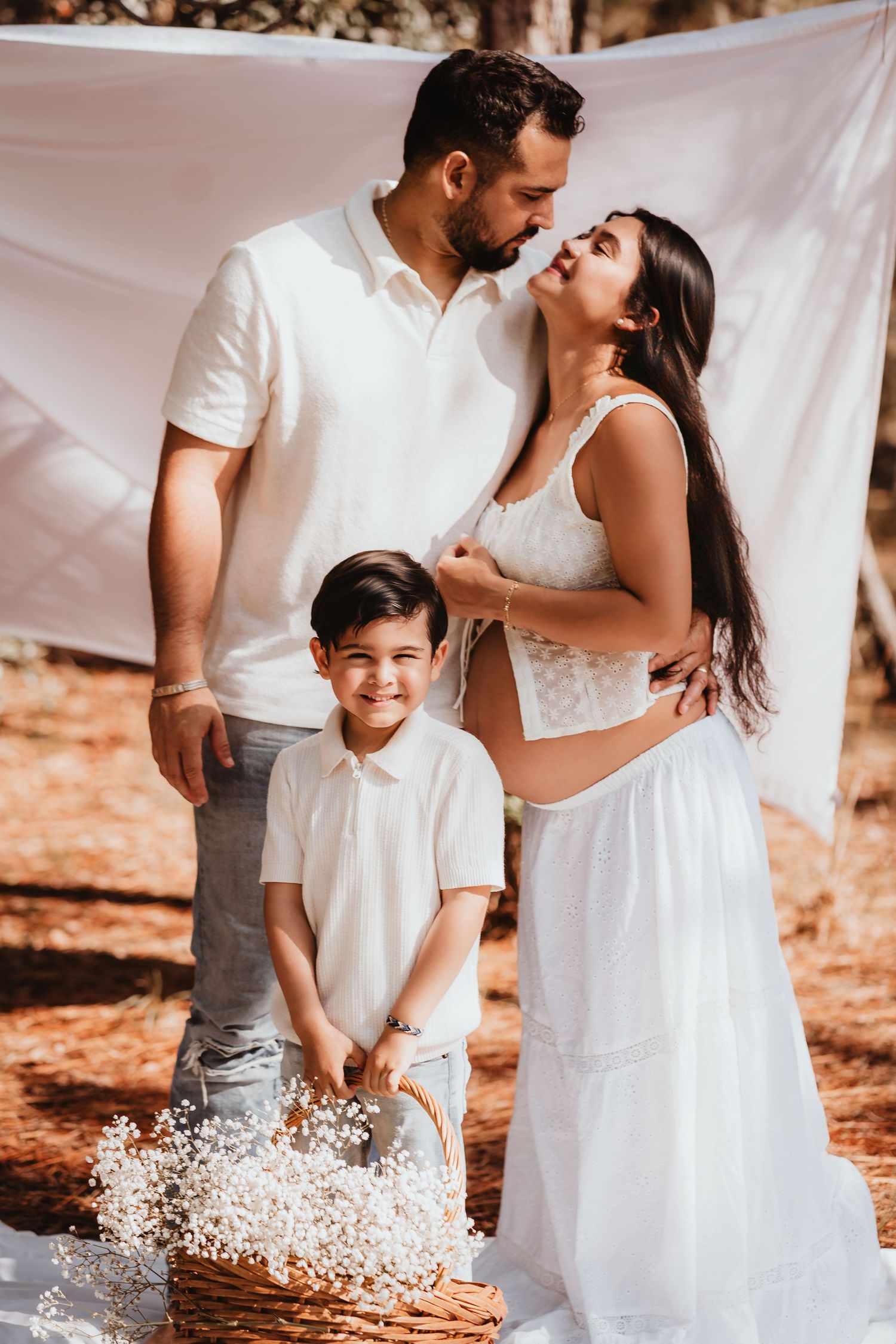 Couple sharing a kiss while young son holds flower basket during outdoor maternity shoot.