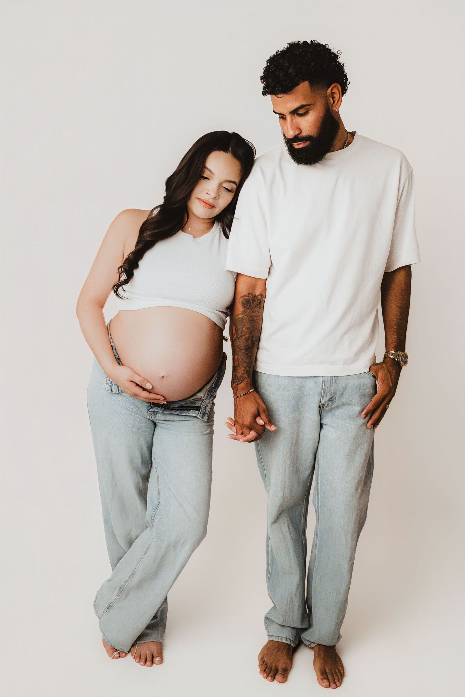 Pregnant woman in white crop top holding belly while partner with tattoos holds her hand tenderly.