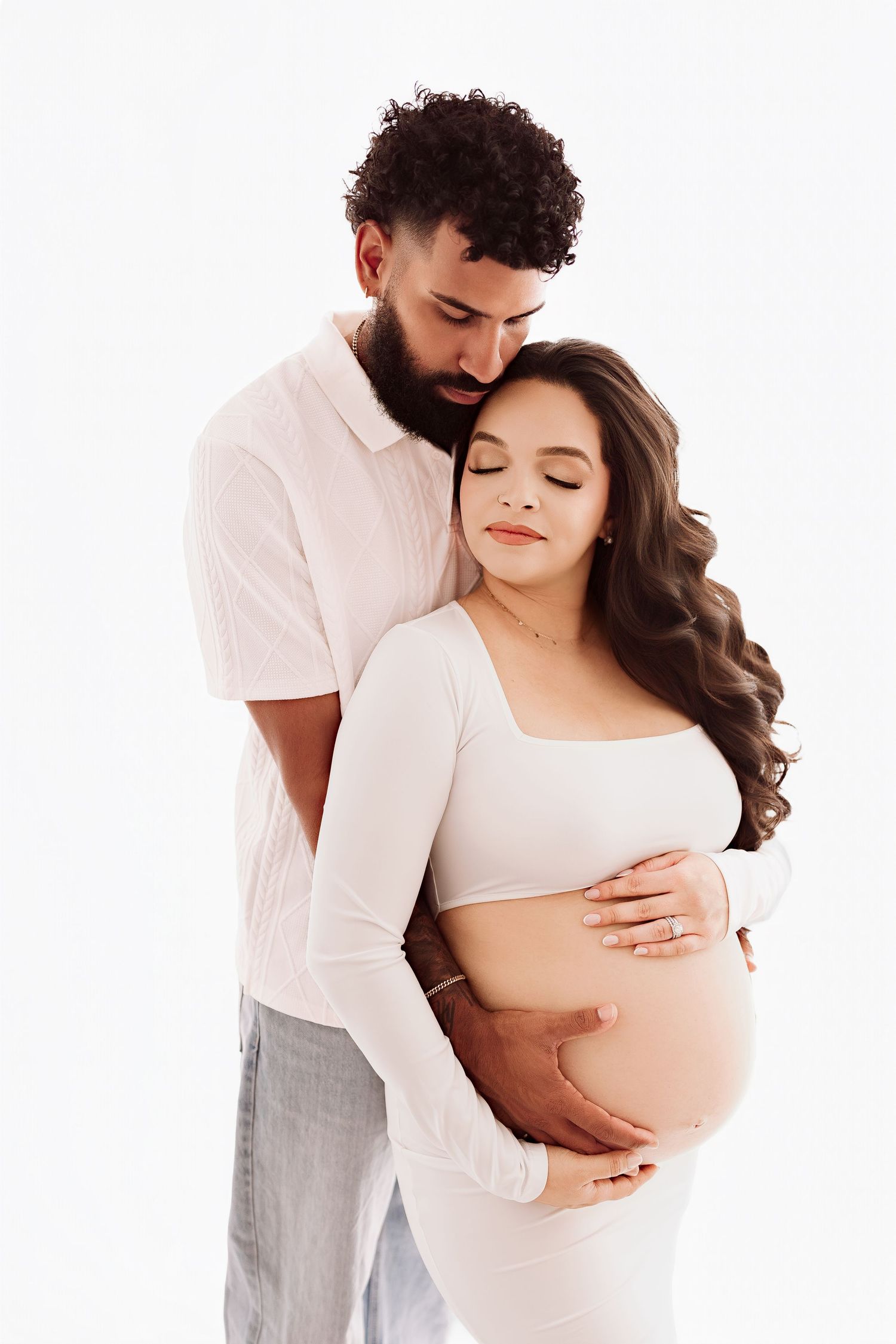 Loving couple in maternity photoshoot, man embracing pregnant woman from behind as both cradle her bare baby bump.