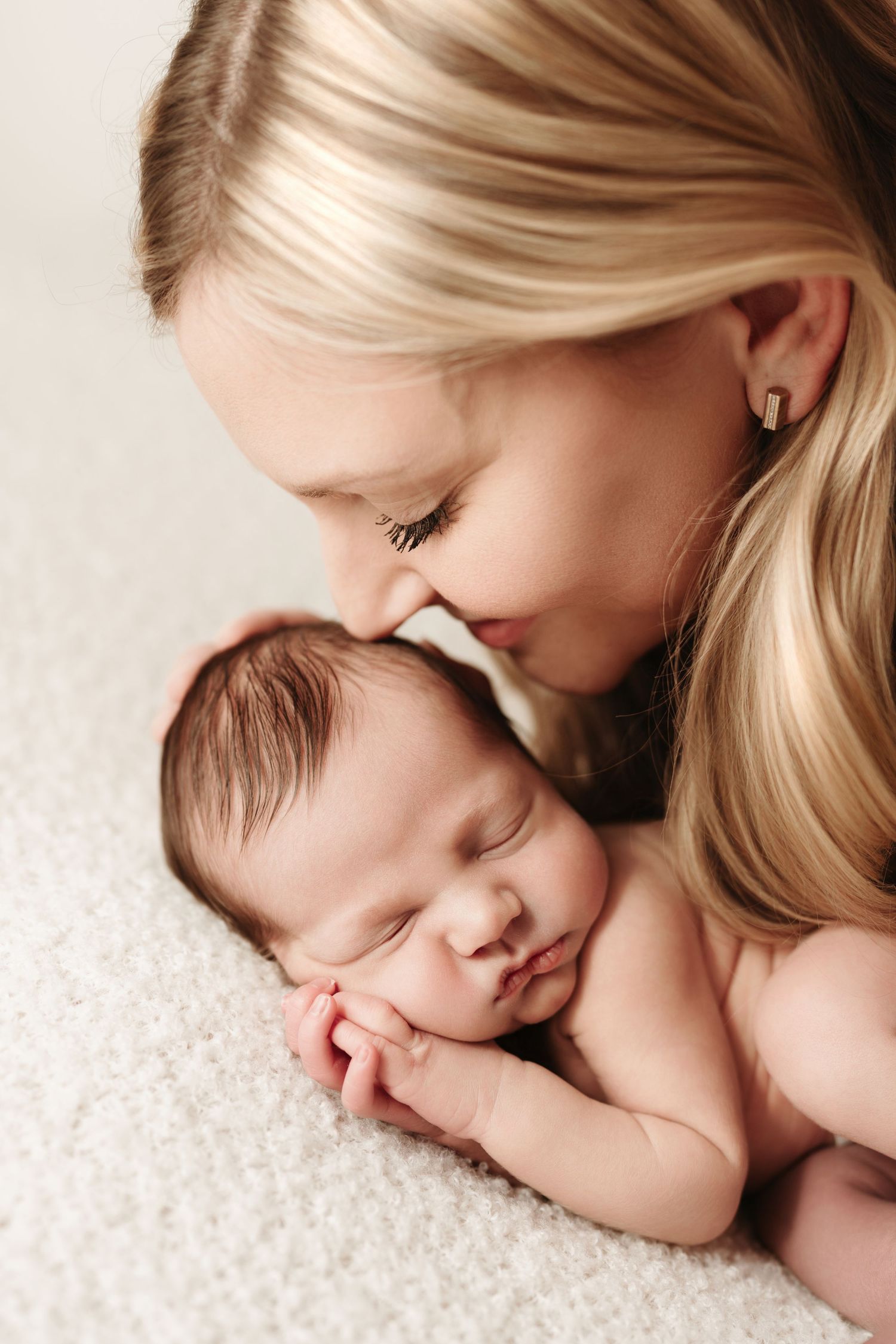 Loving mother with long blonde hair gently kissing sleeping newborn baby's head.
