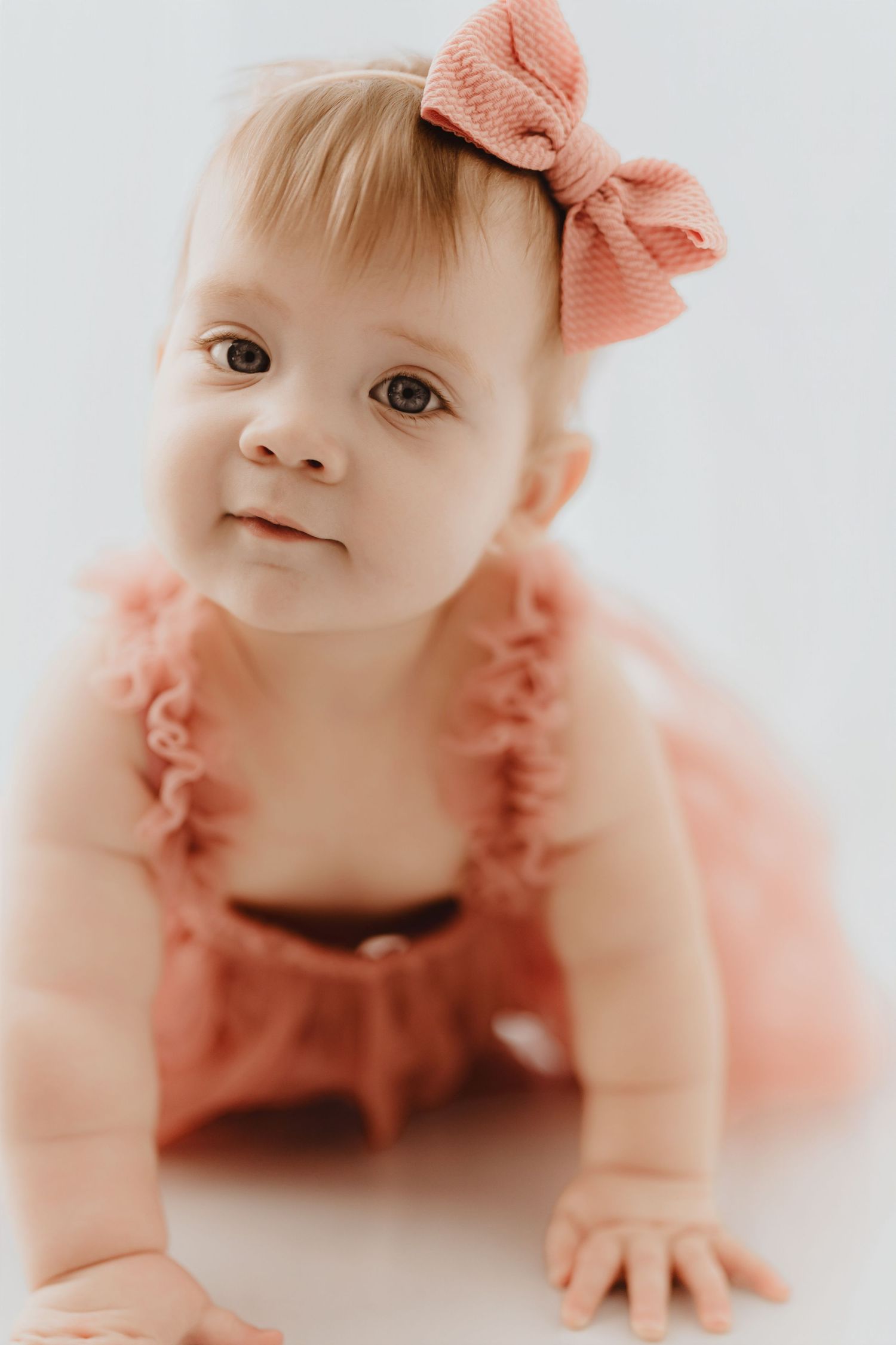 Close-up of baby girl in pink ruffled romper with bow headband crawling on white studio surface.
