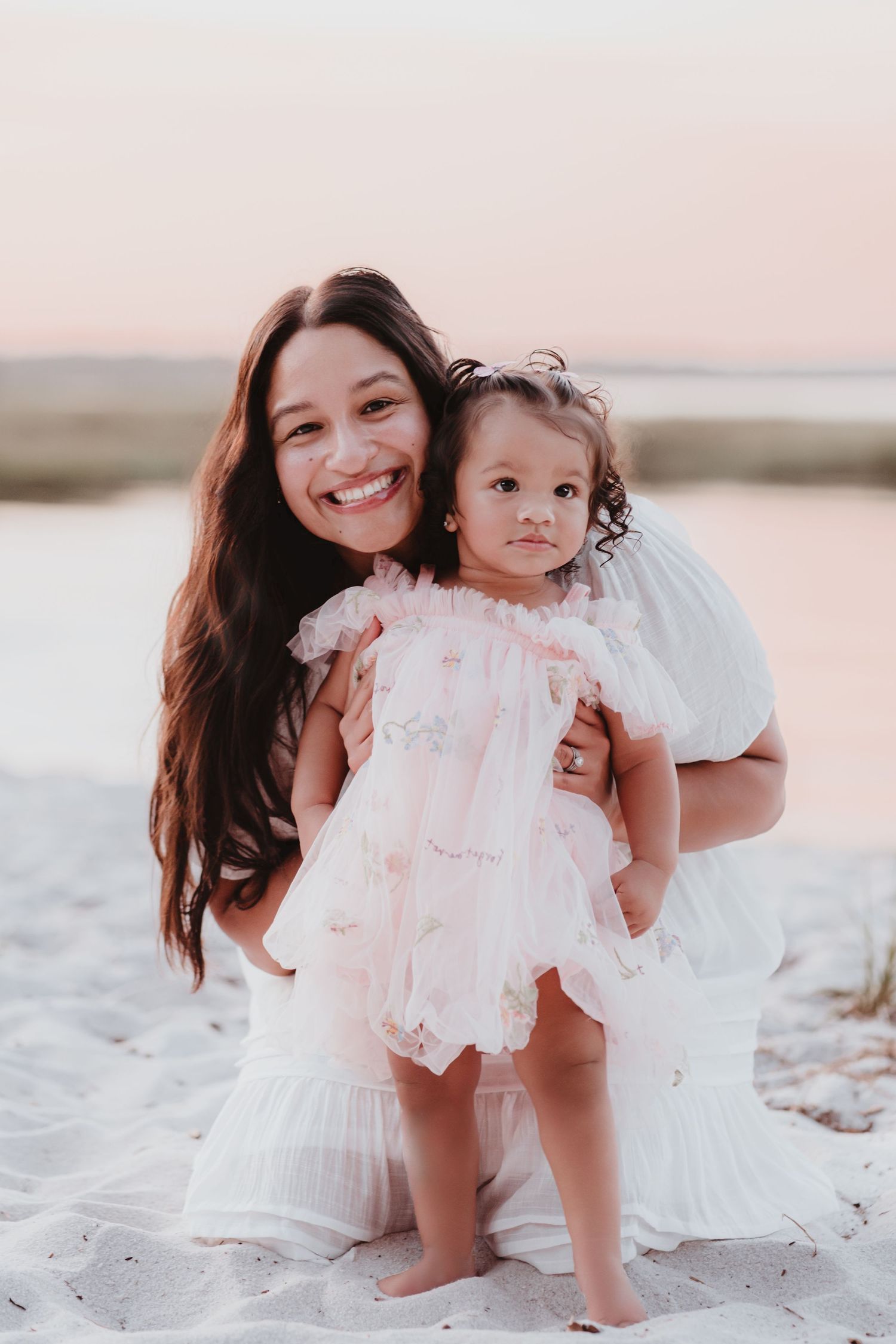 Smiling mother in white dress posing with toddler daughter on sandy beach.
