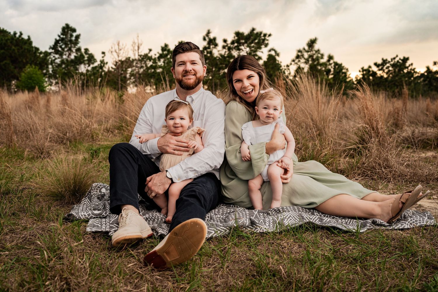 Smiling family of four with two baby girls sitting together on a blanket in a golden sunset field.
