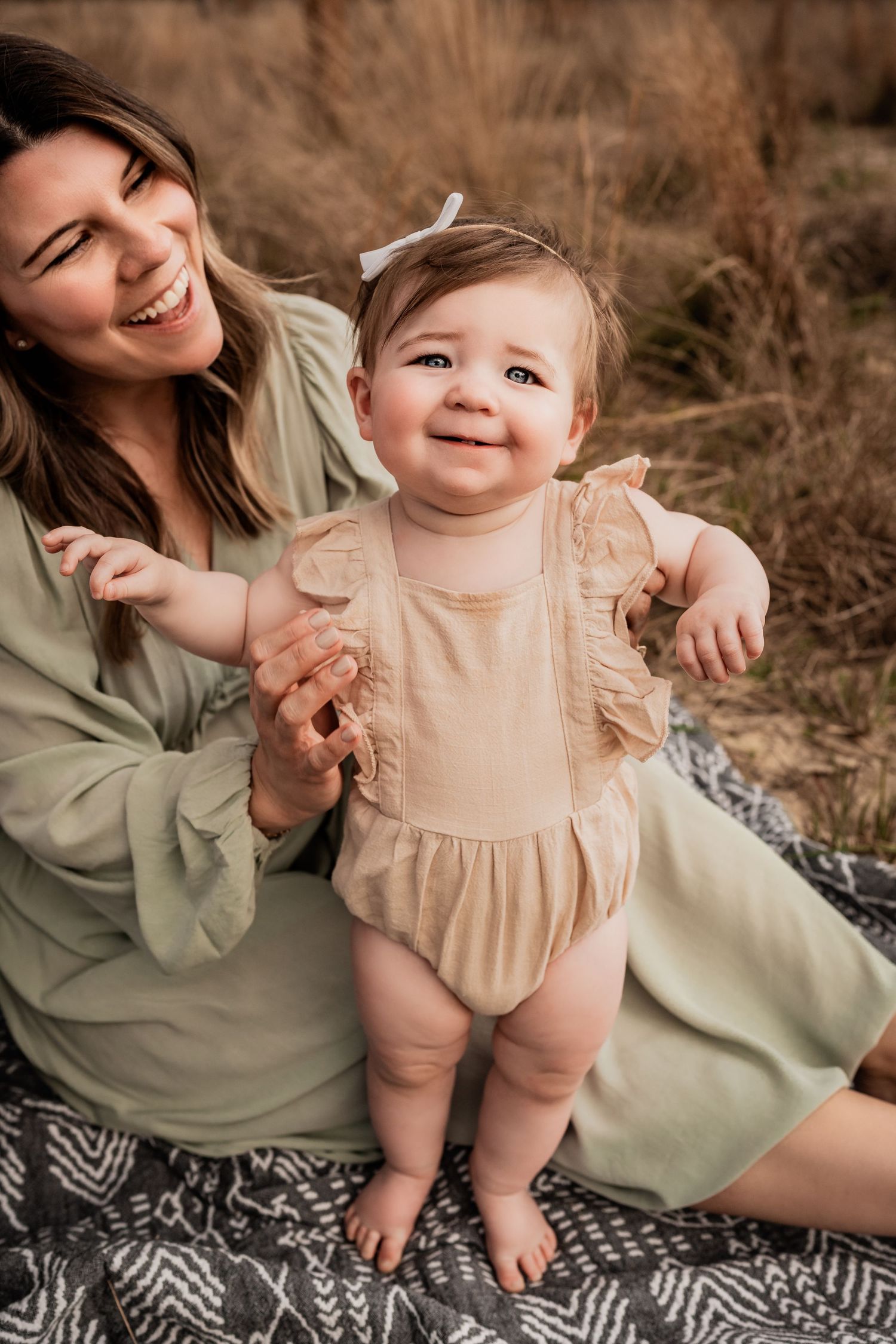 Smiling baby girl in beige ruffle romper standing with support from laughing mother in green dress outdoors.