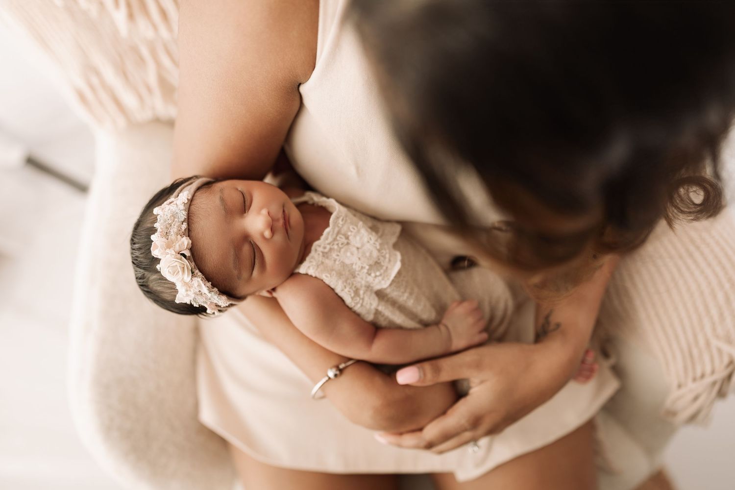 Mother tenderly holding her sleeping newborn baby dressed in a delicate lace outfit with a floral headband.