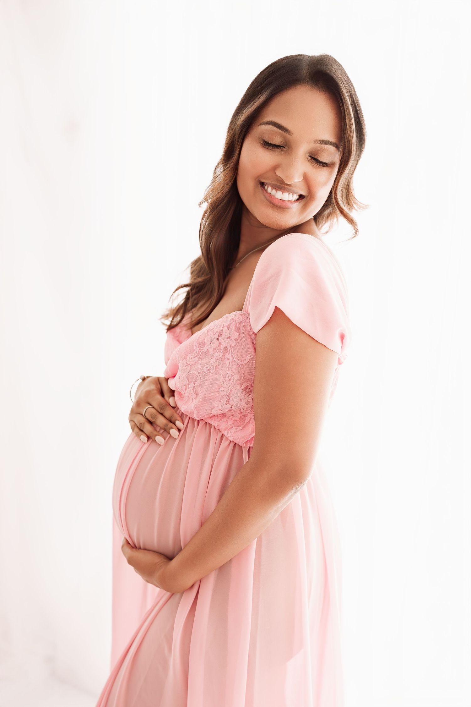 Smiling pregnant woman in pink lace maternity dress gently holding her baby bump.
