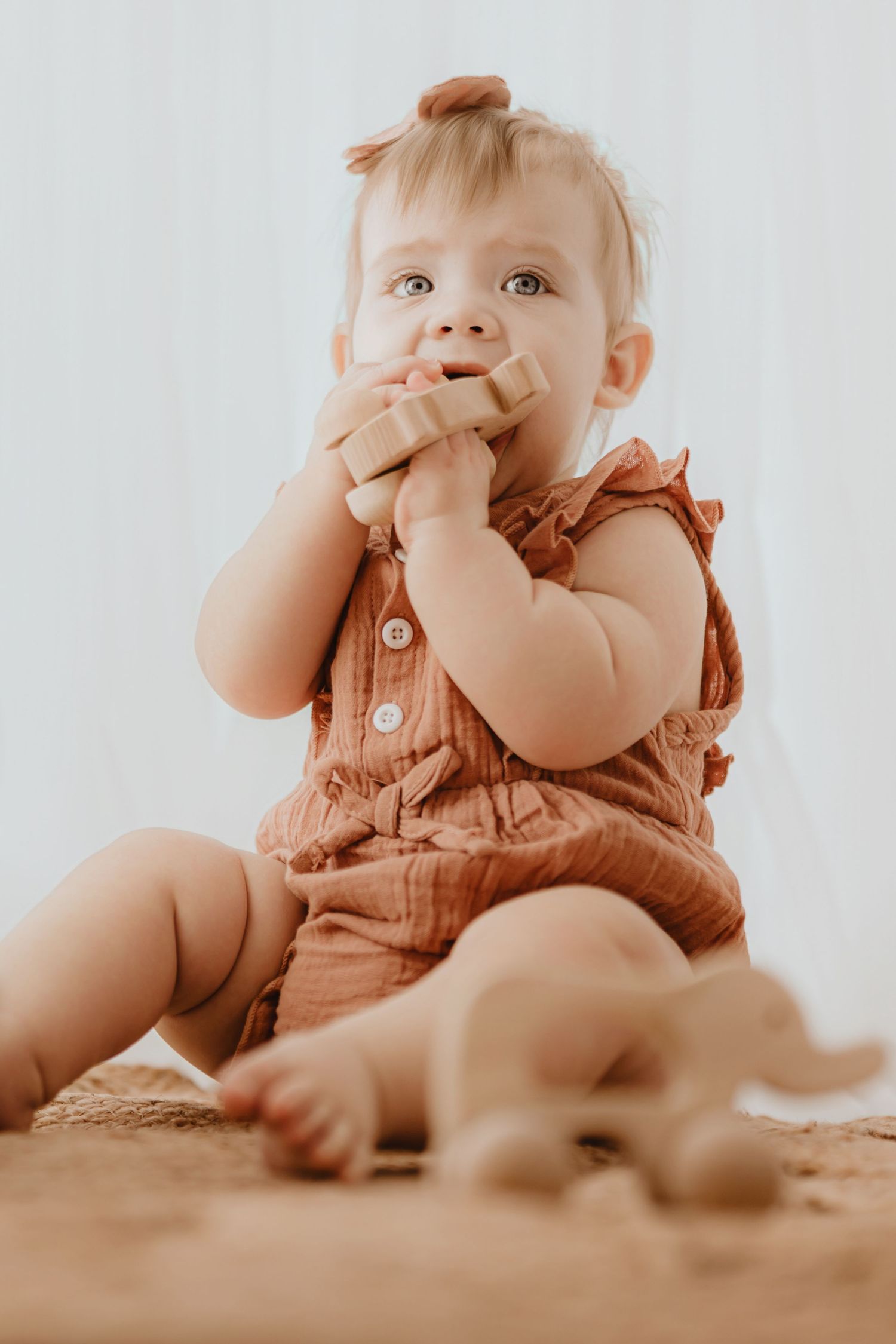 Baby girl in terracotta ruffle romper chewing on wooden toy while sitting on woven rug.