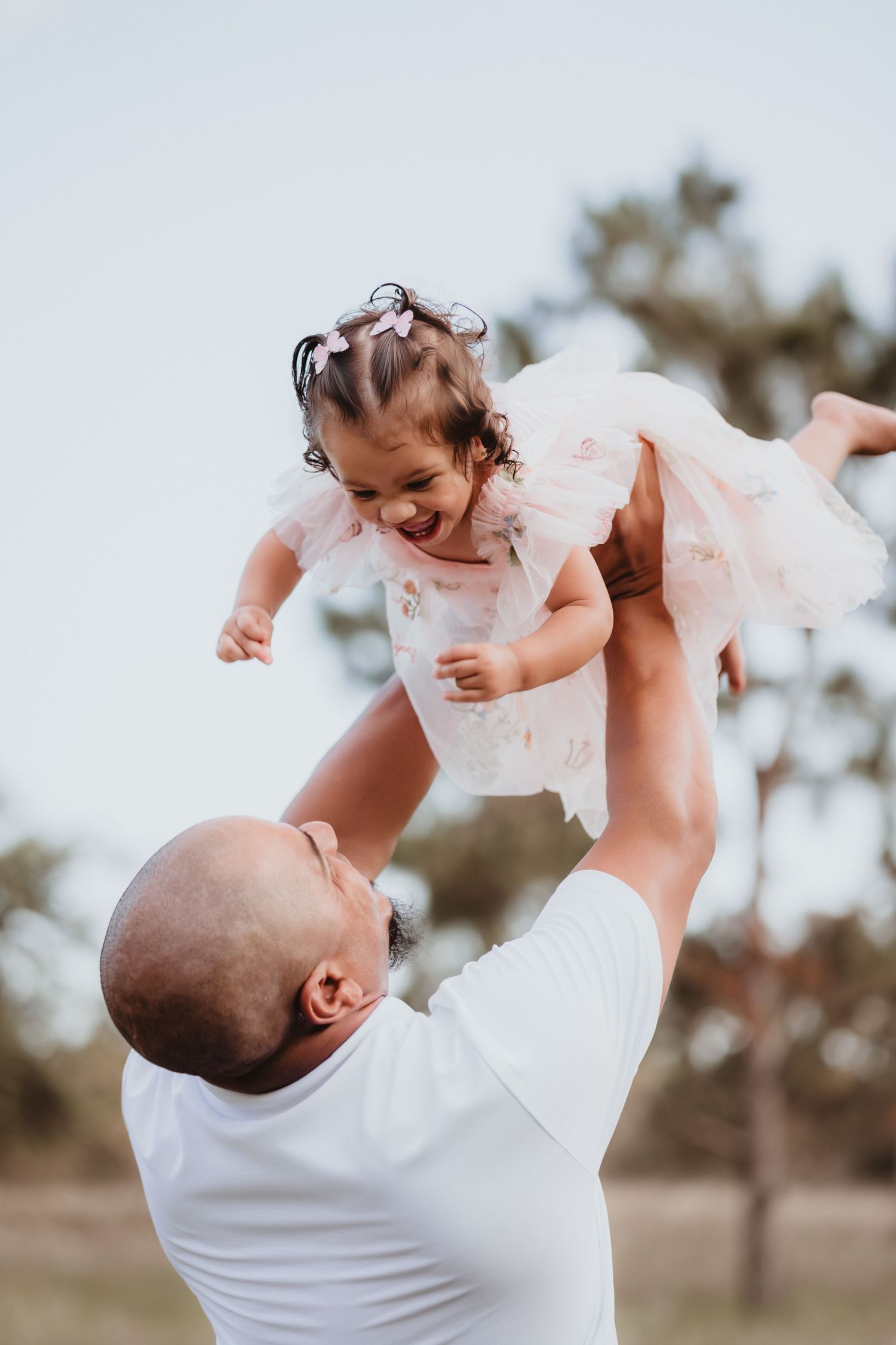 Father joyfully lifting his laughing toddler daughter in a pink tulle dress outdoors.