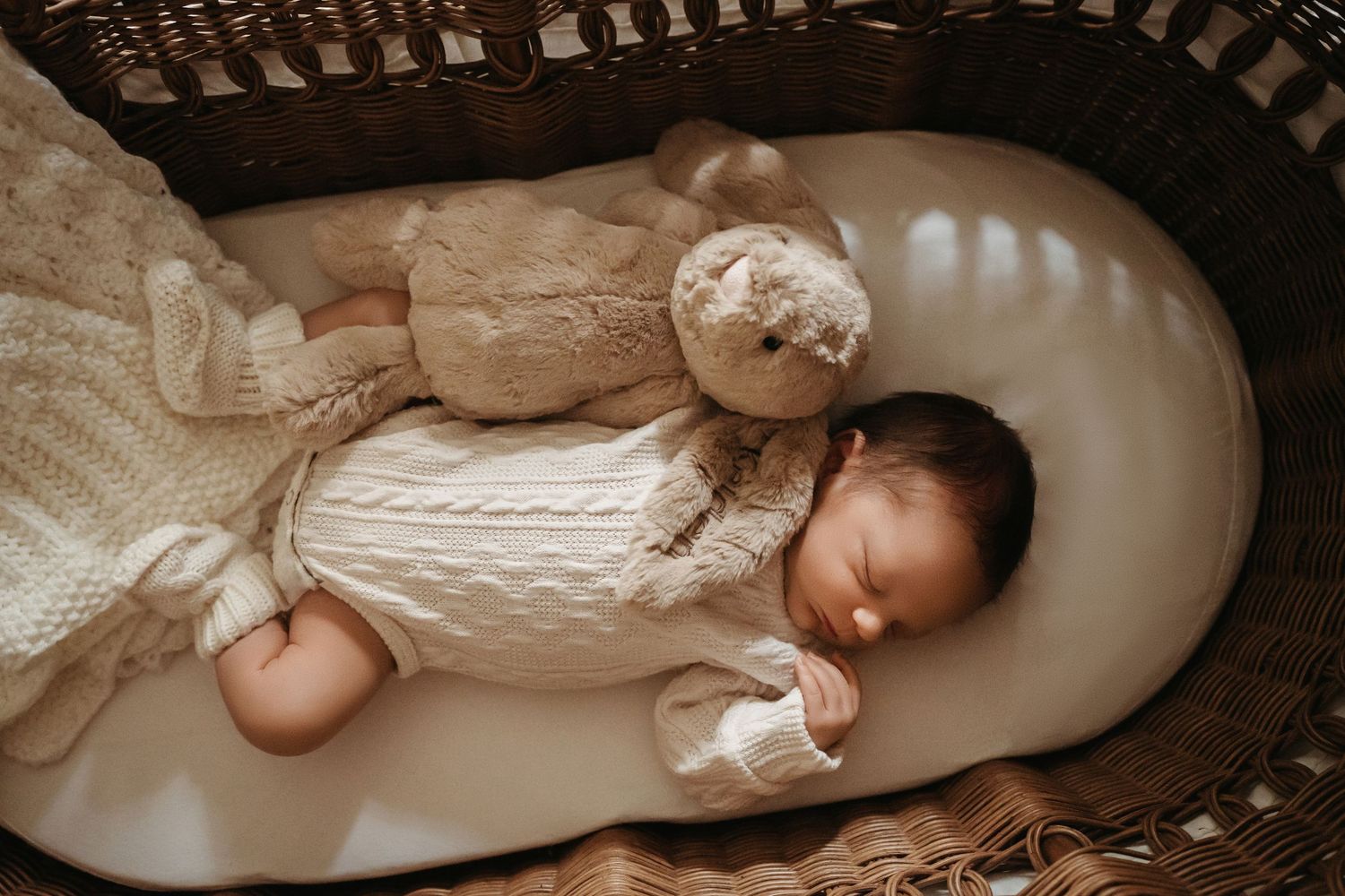 Sleeping newborn baby in white knit outfit resting beside teddy bear in wicker bassinet.