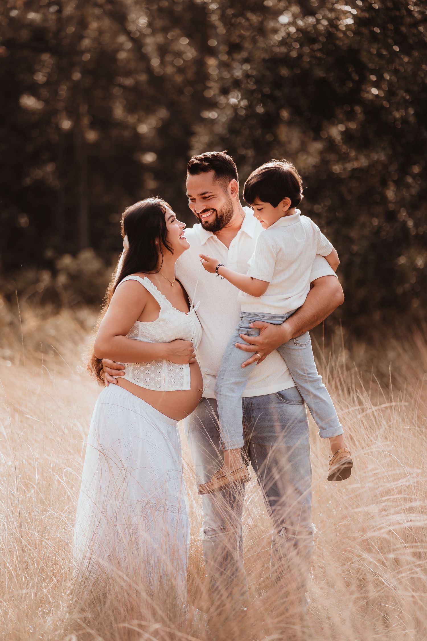 Happy family maternity photo shoot in golden field, pregnant woman in white dress with husband holding young son.