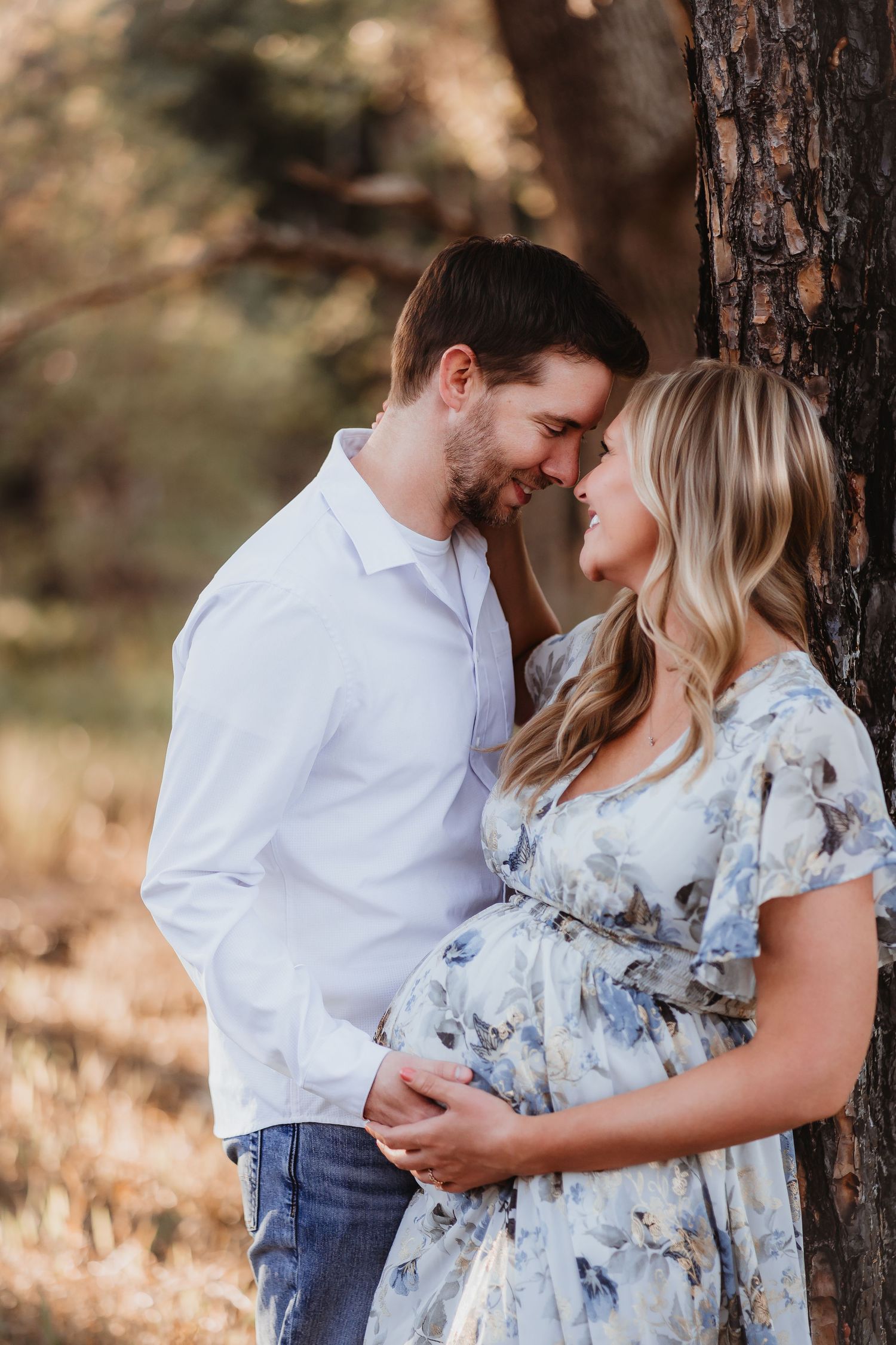 Couple leaning foreheads together smiling near tree during outdoor maternity photo session.