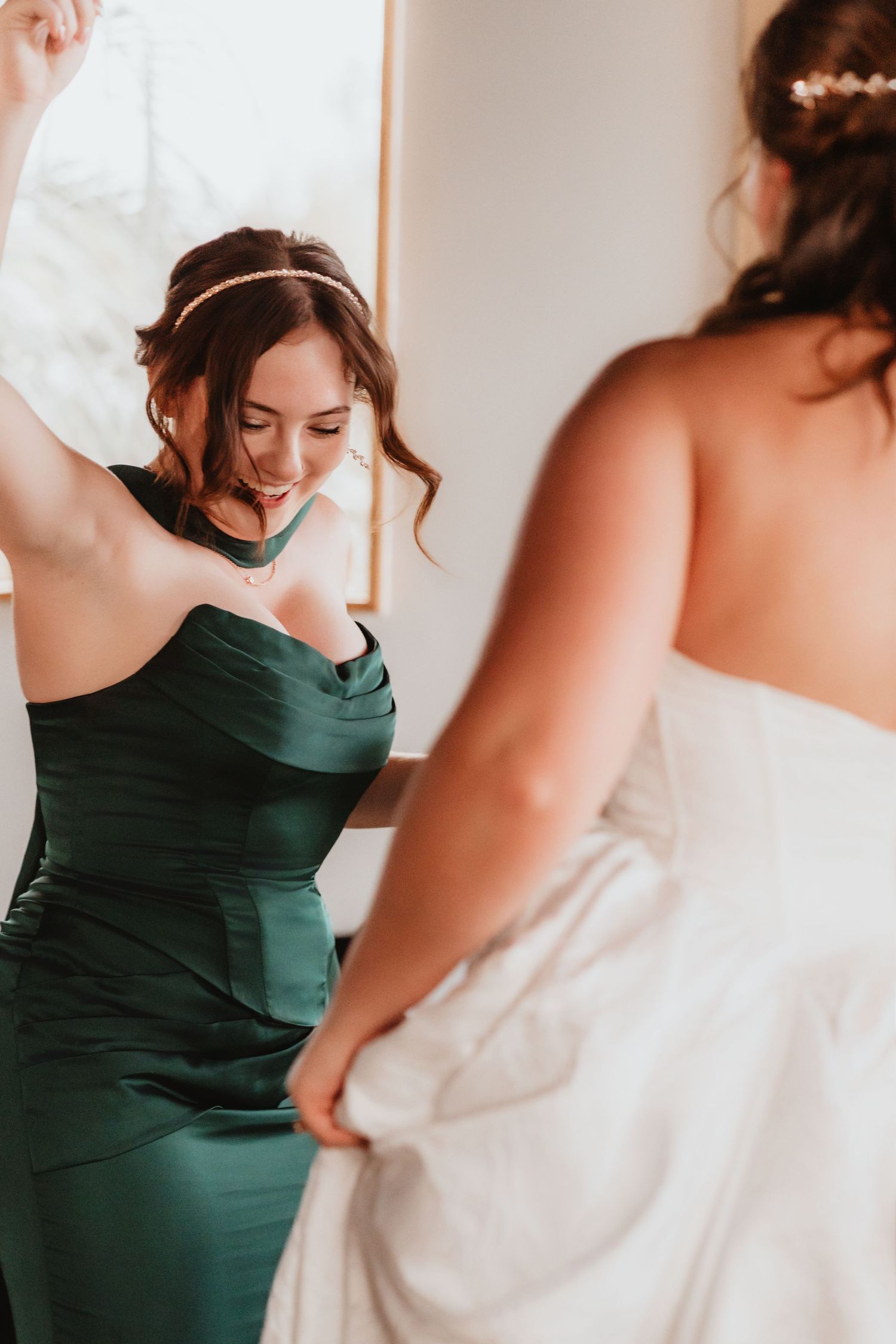 Bridesmaid in emerald green gown helps bride with white wedding dress during bridal preparations.