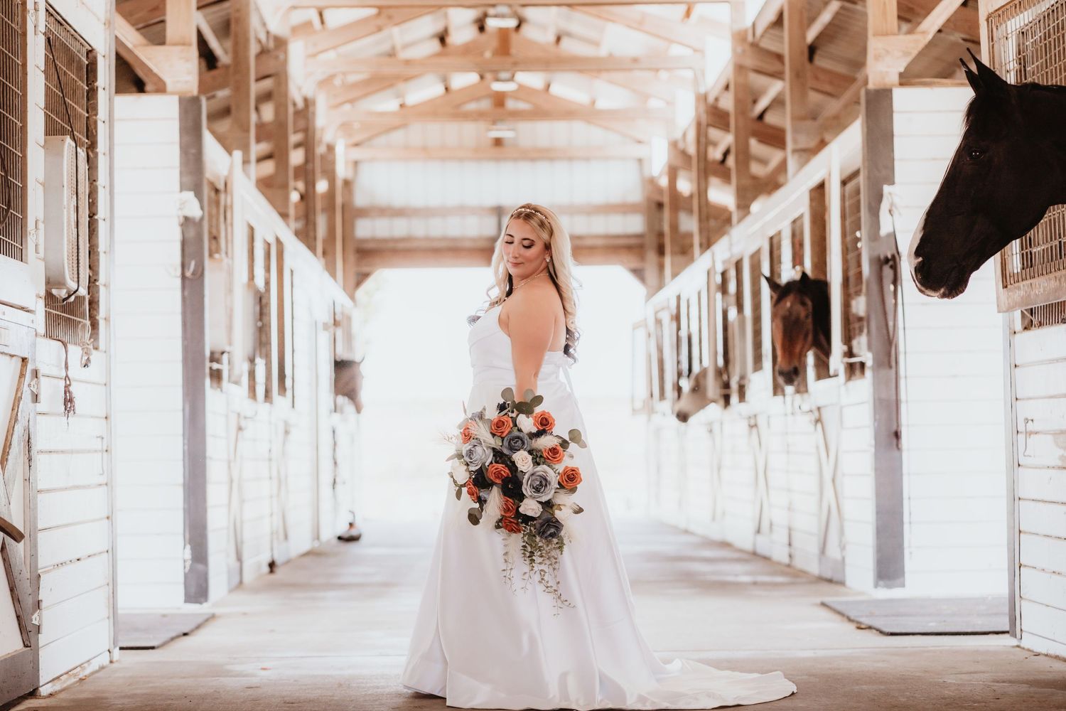 Bride in a flowing white gown walks through a bright horse stable holding a colorful floral bouquet.