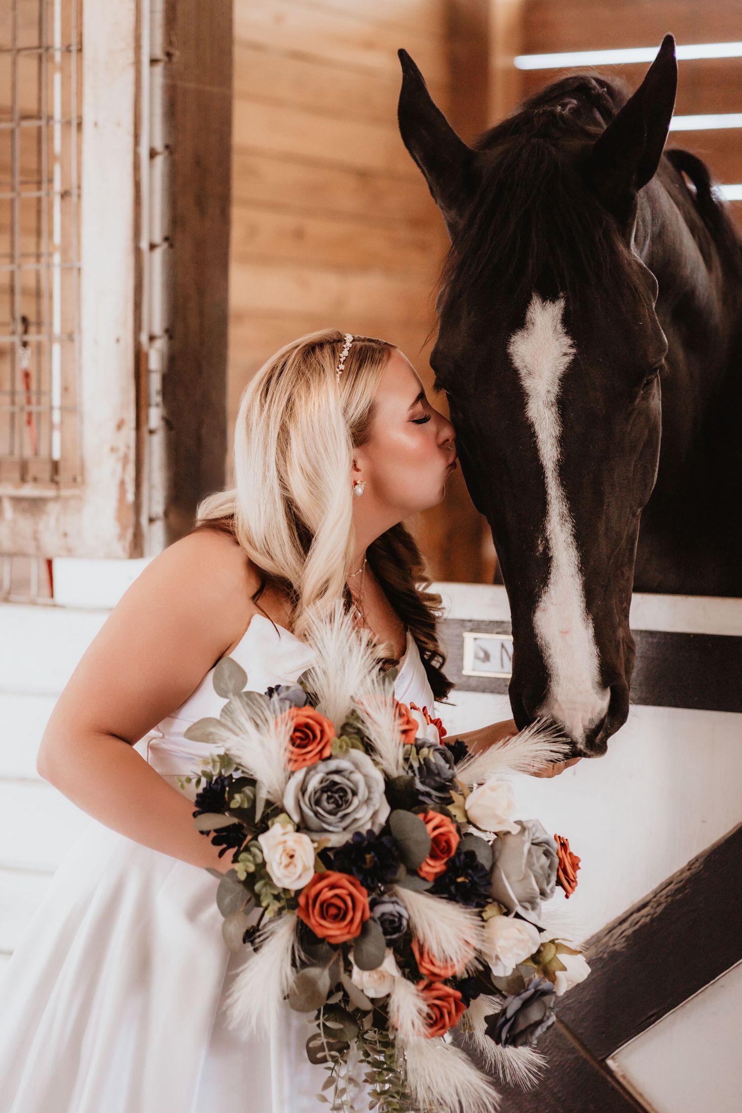 Bride in white gown kisses a dark horse in a stable while holding a bouquet of orange, gray, and white roses.