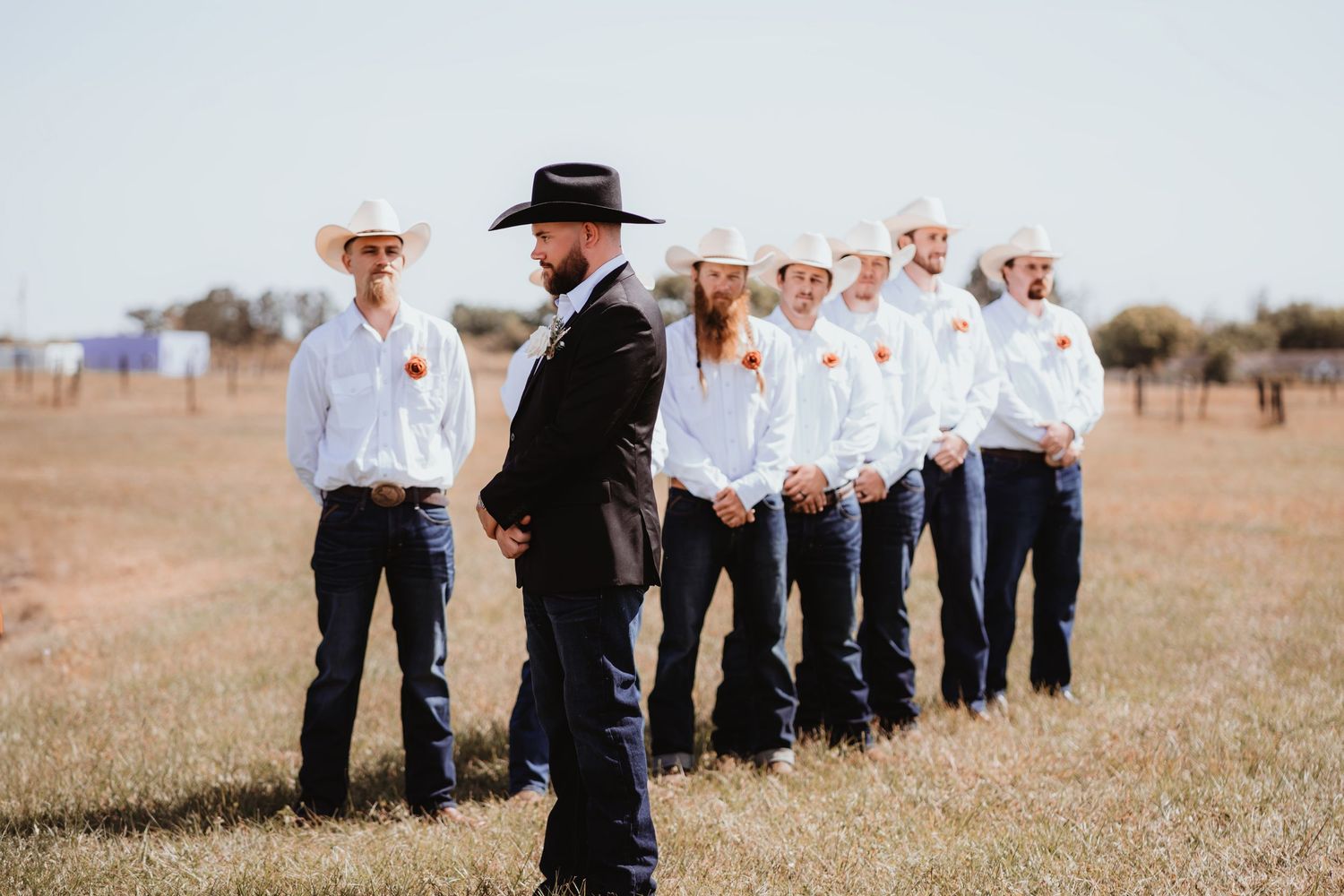 Groom in black hat and suit faces line of groomsmen in white shirts and cowboy hats at outdoor wedding.