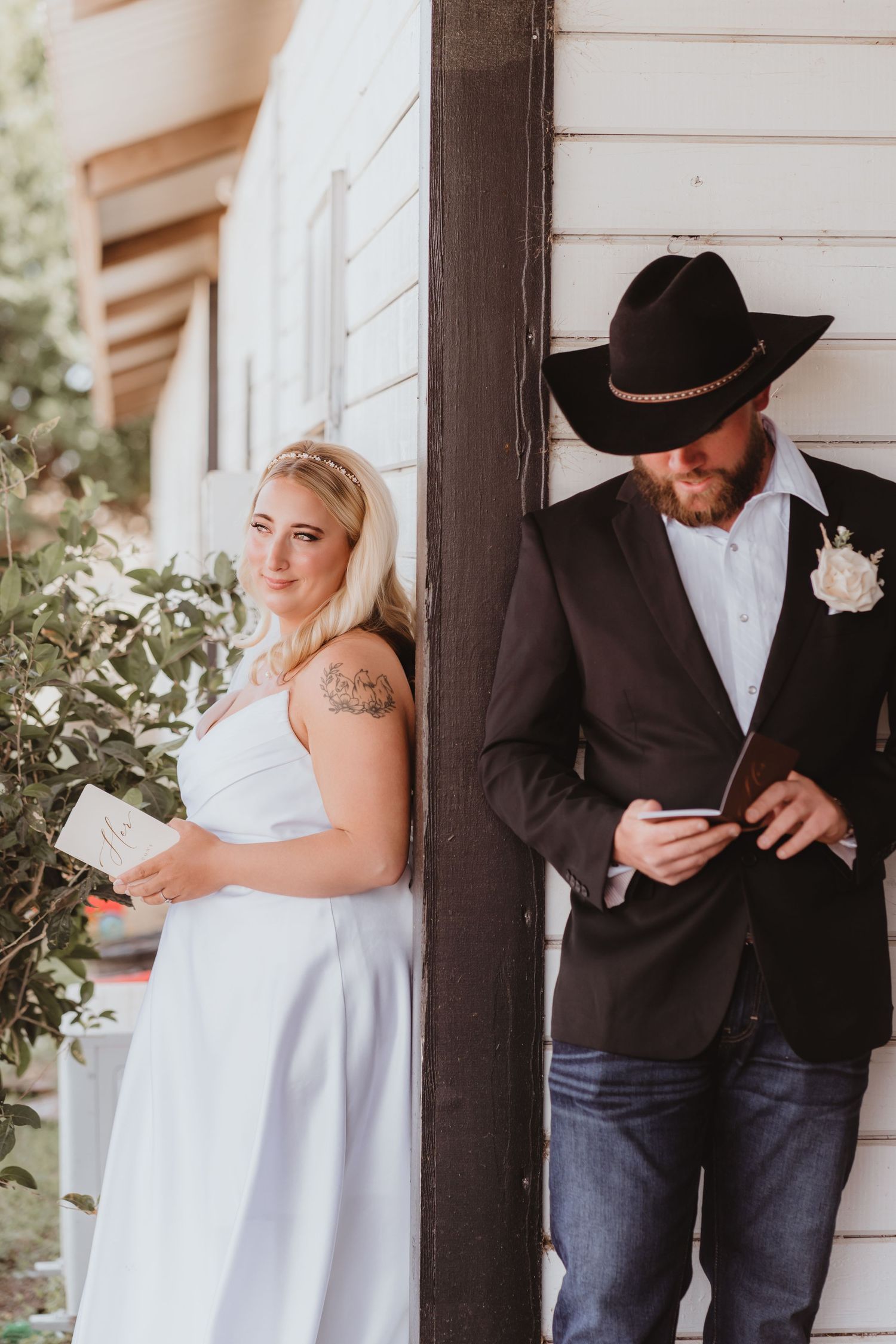 Bride and groom share private vow reading moment leaning against white wooden building exterior.