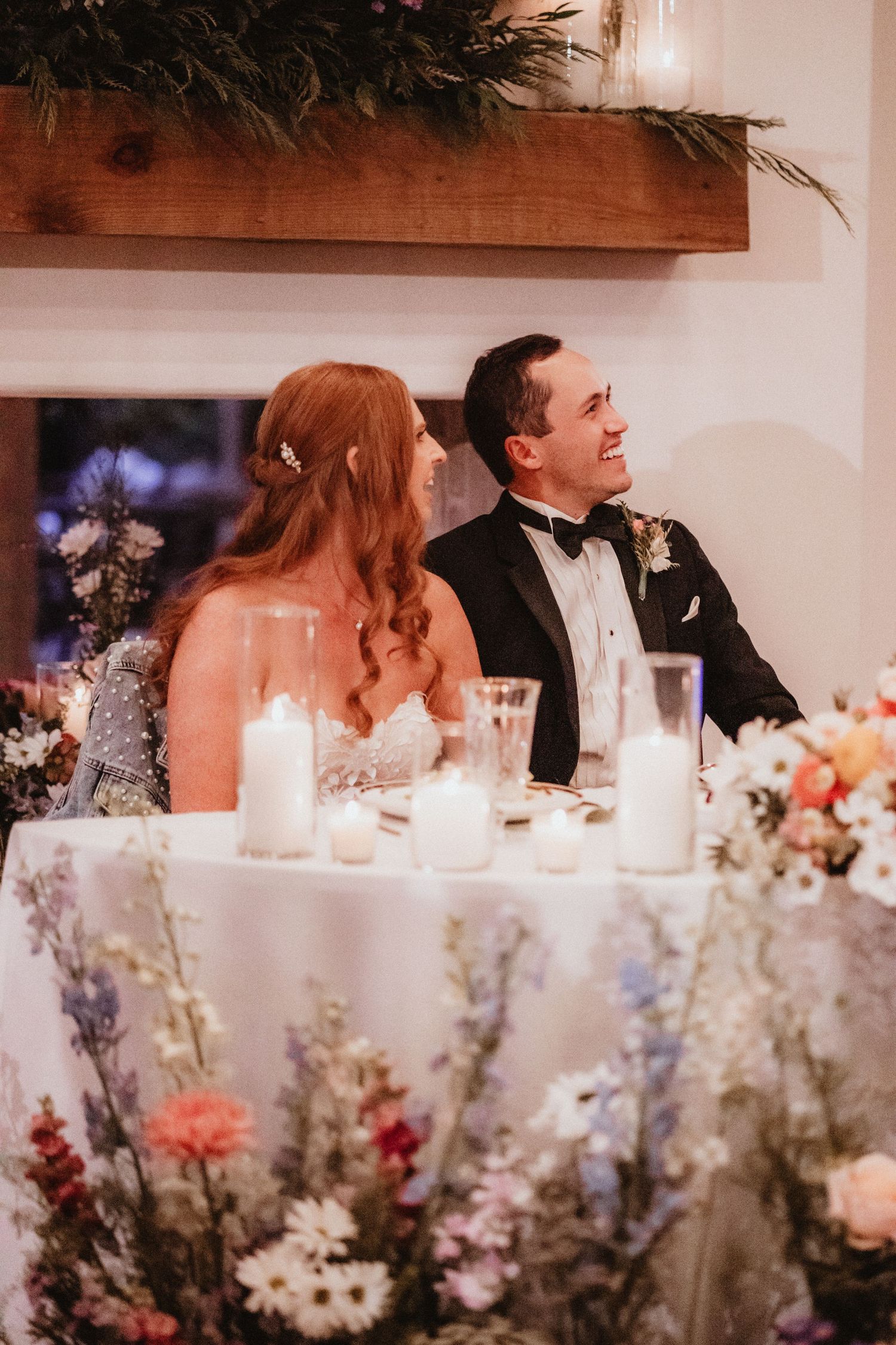 Bride and groom smile at sweetheart table adorned with floral tablecloth and candles at wedding reception.