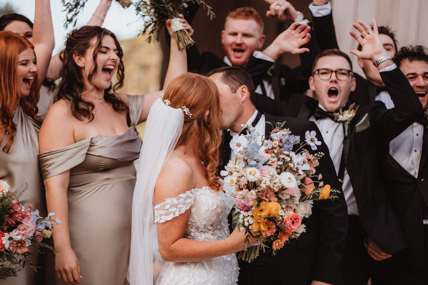 Bride and groom share first kiss surrounded by cheering wedding party in elegant attire.