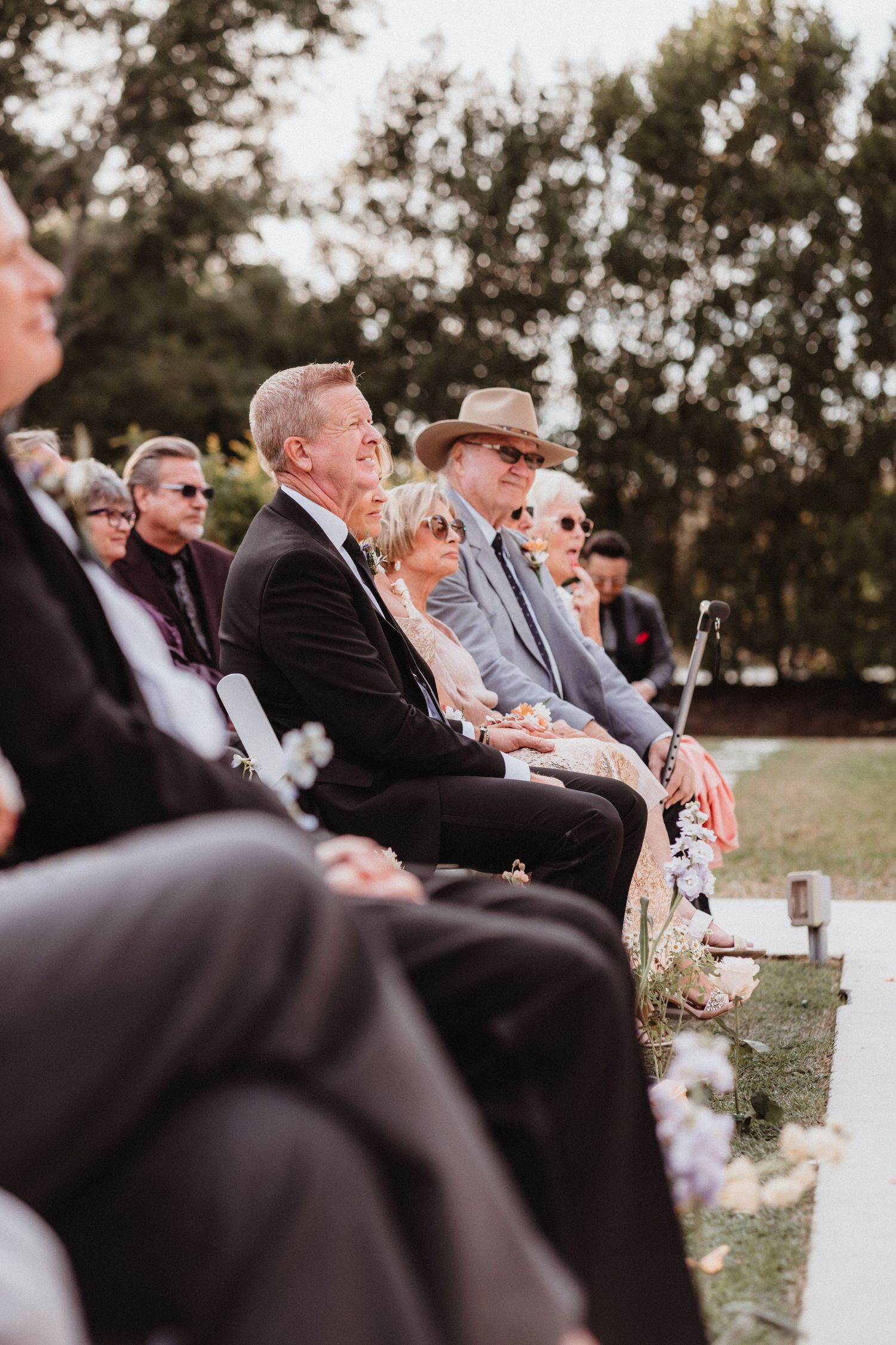 Wedding guests seated outdoors watching ceremony, man in black suit and man in tan hat in foreground.