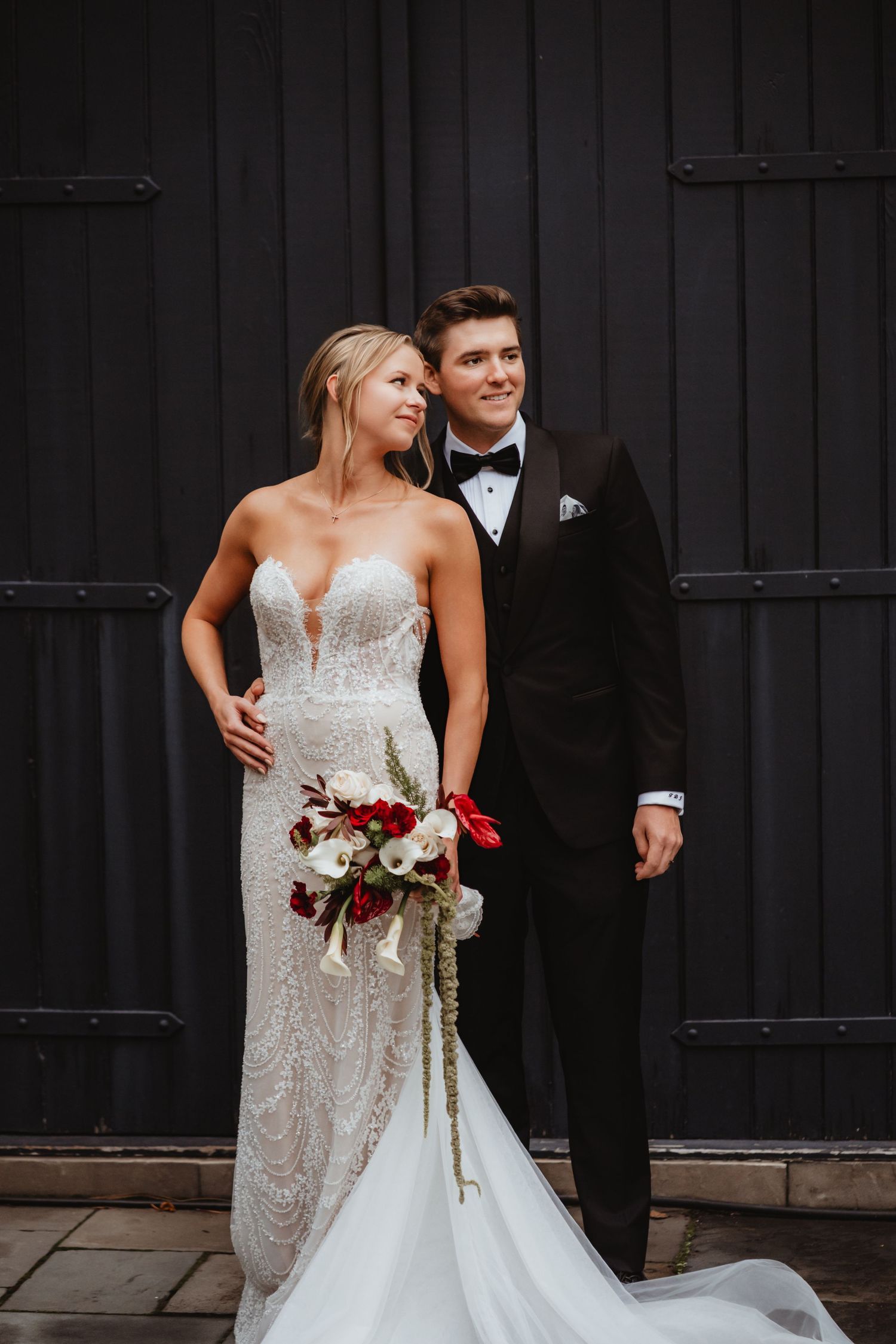 Elegant bride and groom stand before dark barn doors, she in a beaded strapless gown holding a cascading bouquet.