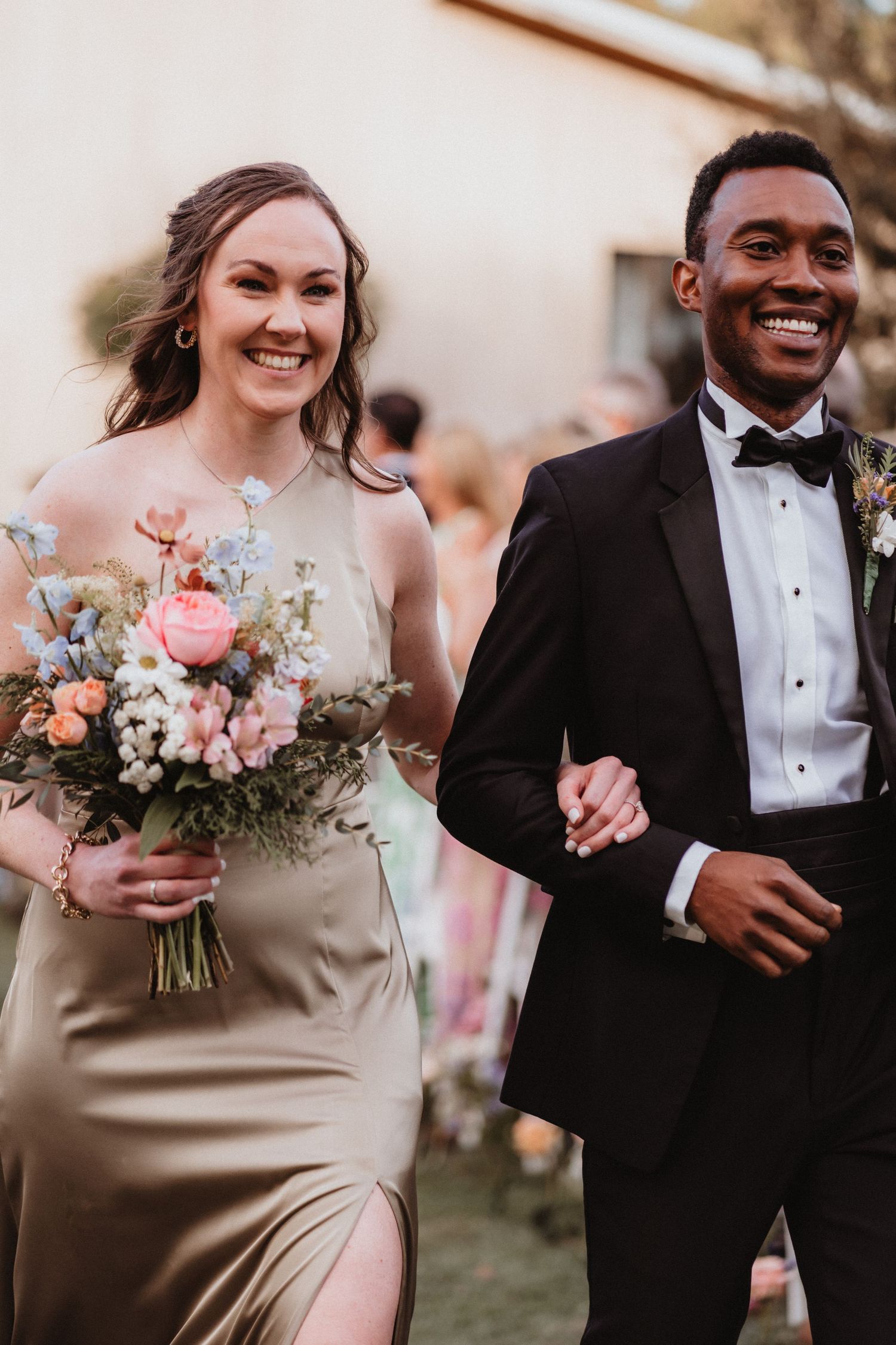 Smiling bridesmaid in gold dress and groomsman in black tuxedo walk together after wedding ceremony.
