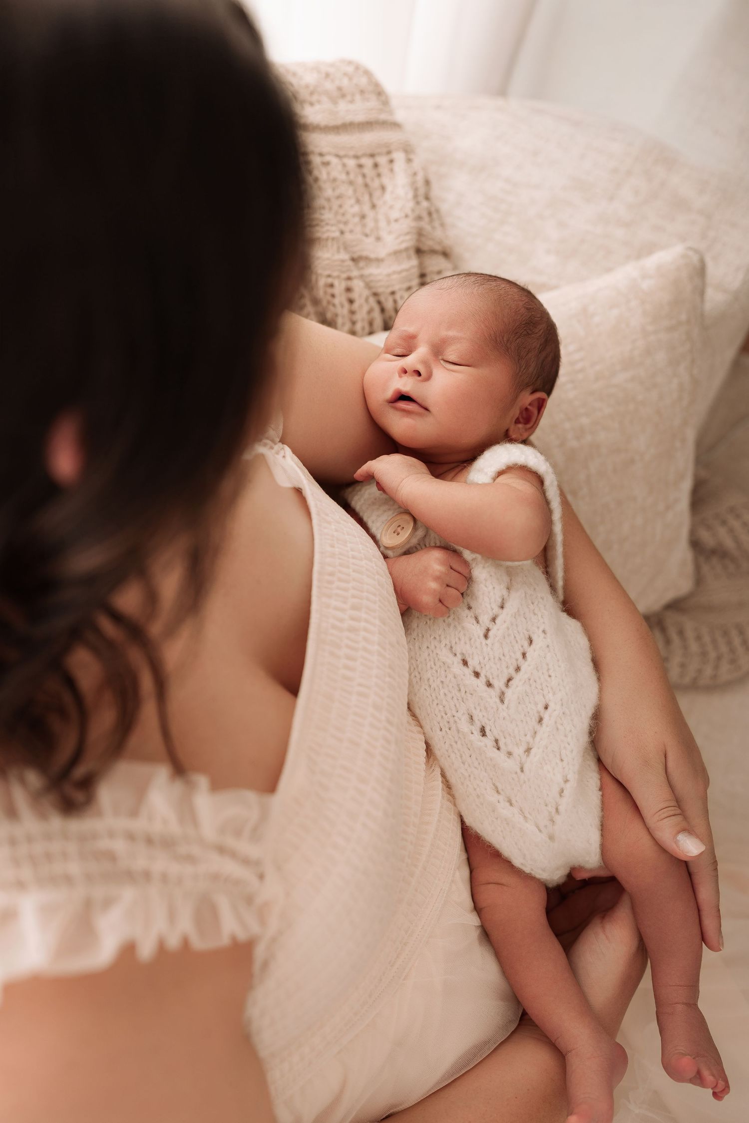 A mother in a white lace dress gently holds her sleeping newborn baby dressed in a cream knit romper.