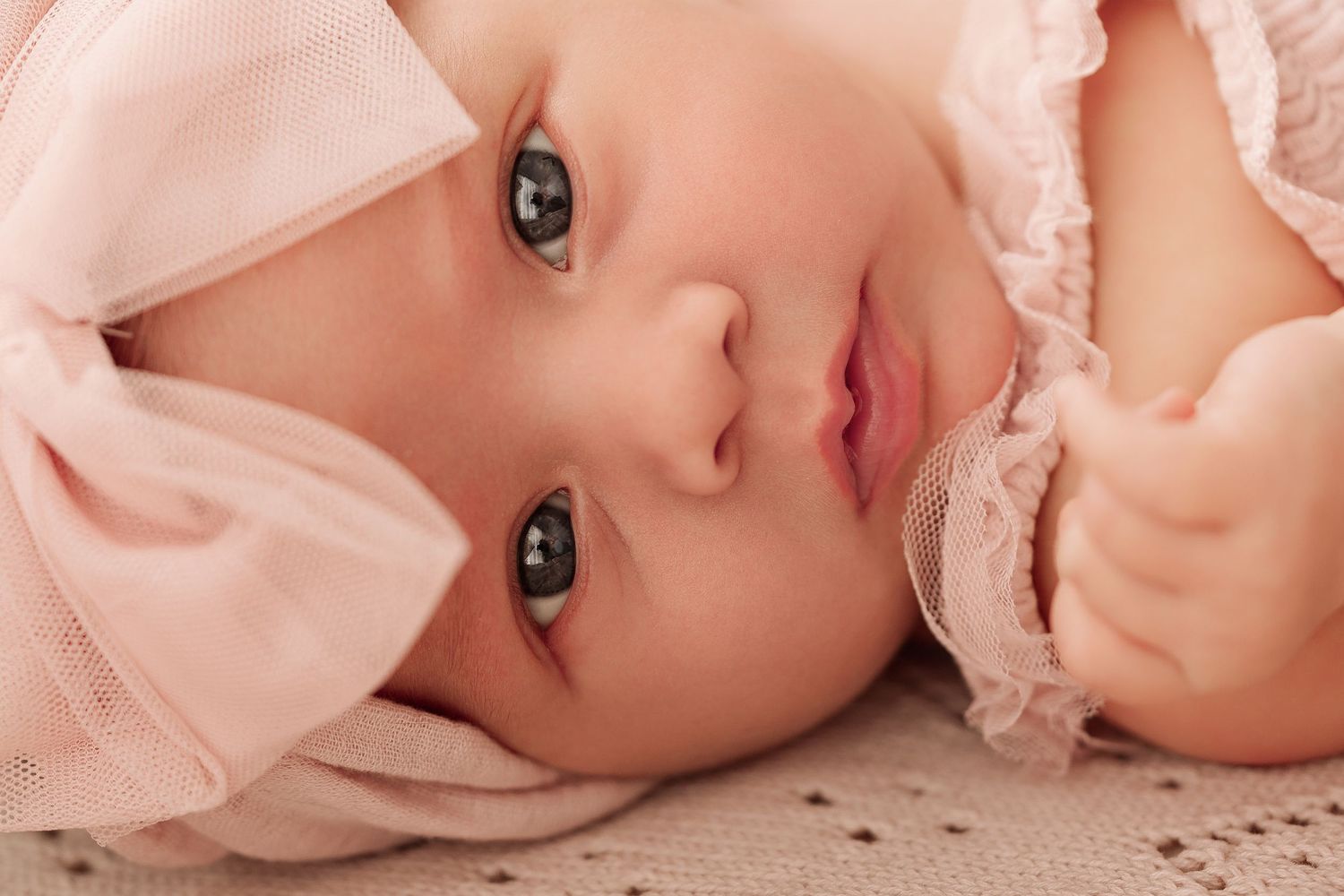 Adorable baby girl in pink outfit with bow headband lying down, looking up with big blue-gray eyes.