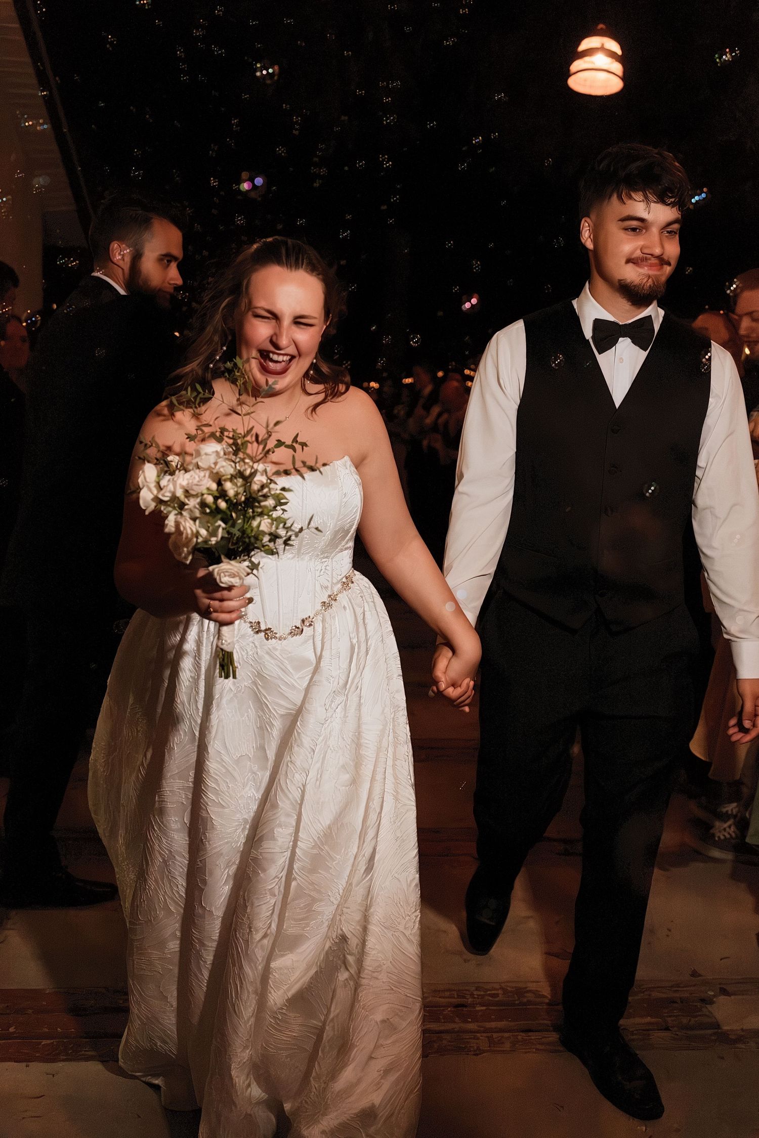 Laughing bride holding bouquet and groom in vest exit wedding reception during joyful nighttime send-off.
