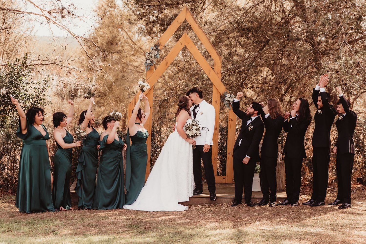 Wedding party with bridesmaids in green and groomsmen cheering under wooden arch at outdoor ceremony.