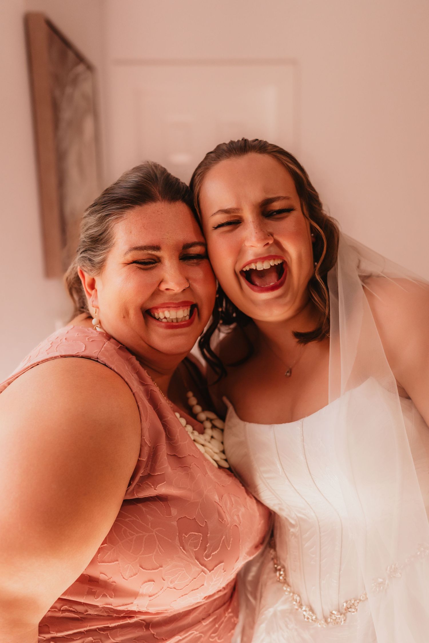 Joyful bride in white gown and veil laughing with bridesmaid in pink dress on wedding day.