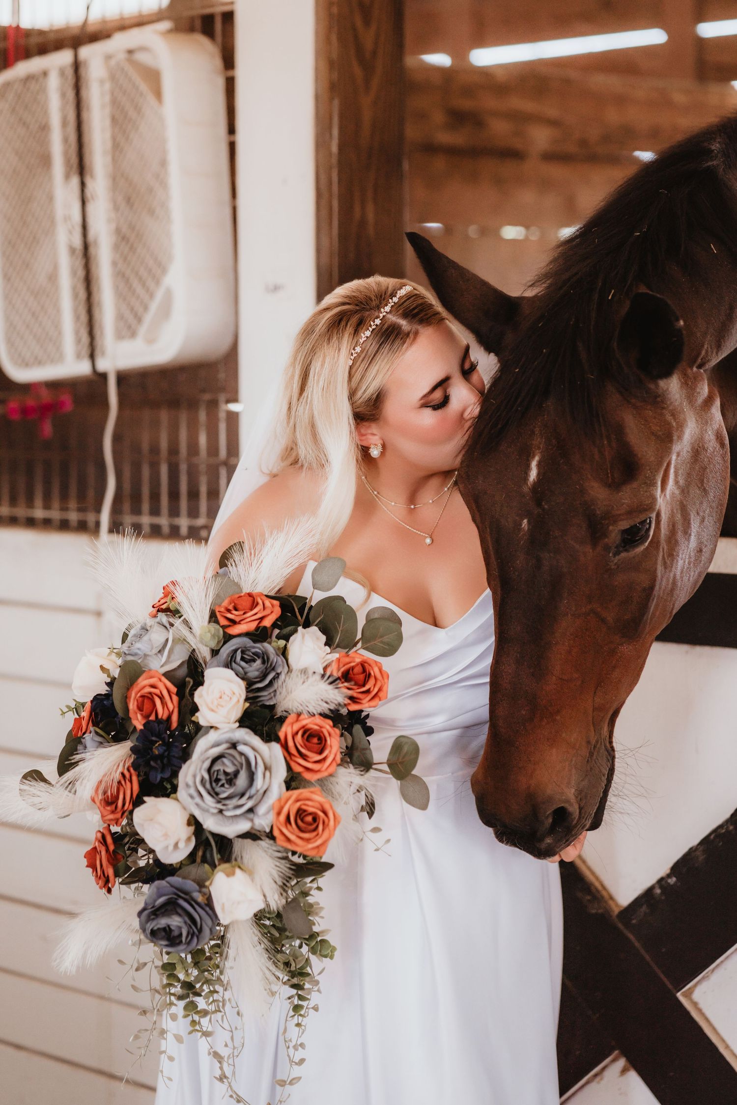 Blonde bride in strapless white dress kisses a brown horse while holding a cascading floral bouquet.