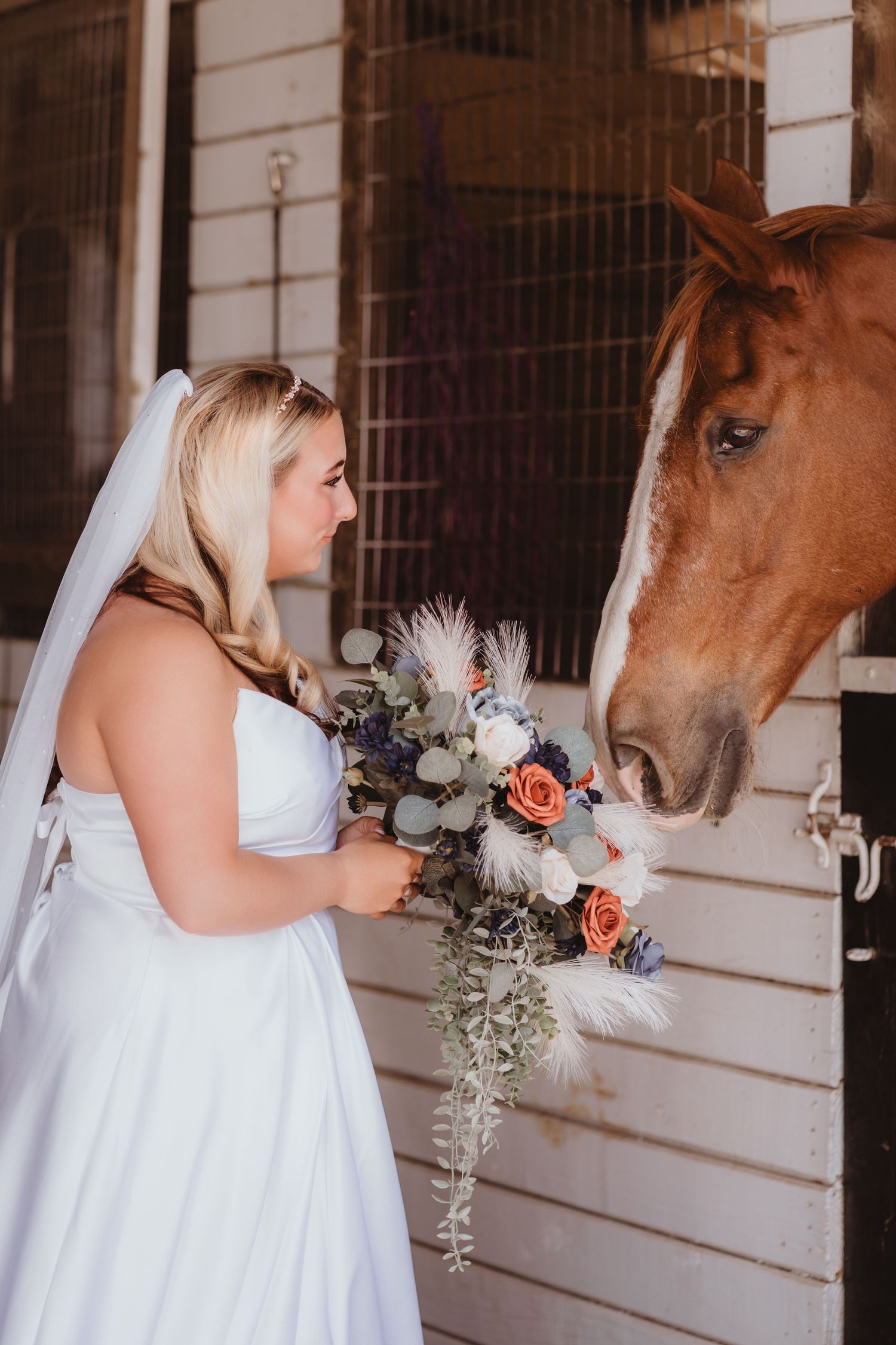 Bride in white gown holds floral bouquet near a chestnut horse in a stable stall.