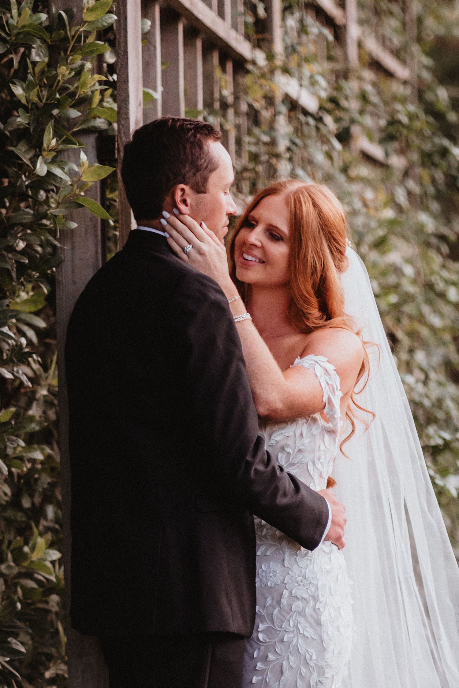 Bride in off-shoulder gown embraces groom tenderly in a garden setting.