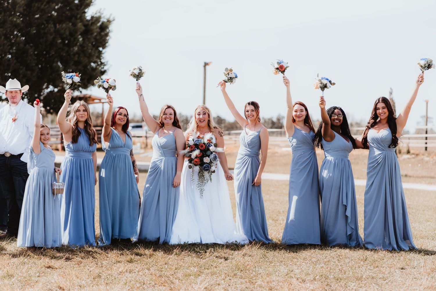 Smiling bride with bridesmaids in dusty blue dresses raising bouquets at an outdoor rustic wedding.