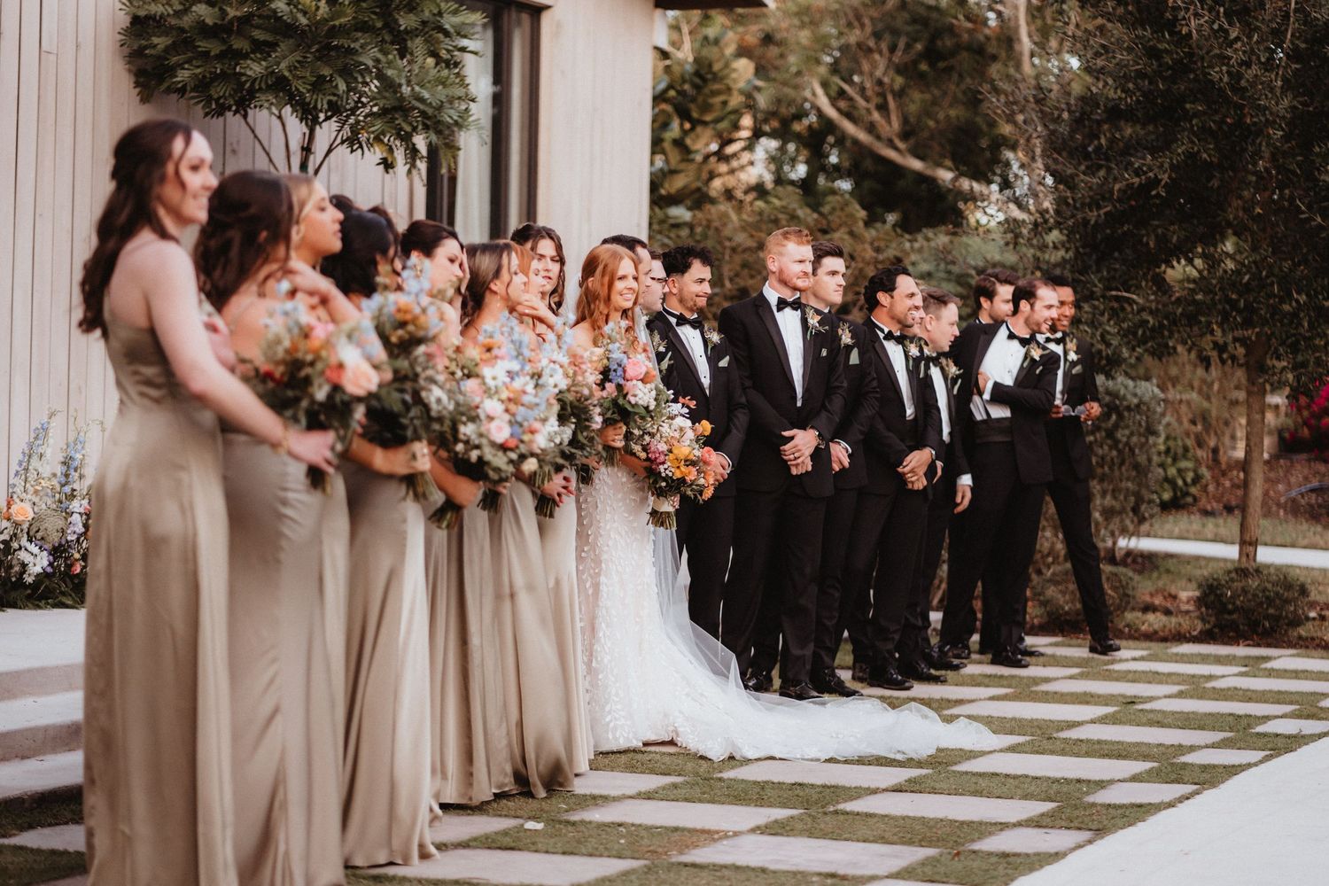 Wedding party lines up outdoors, bridesmaids in taupe dresses holding floral bouquets beside groomsmen in black tuxedos.