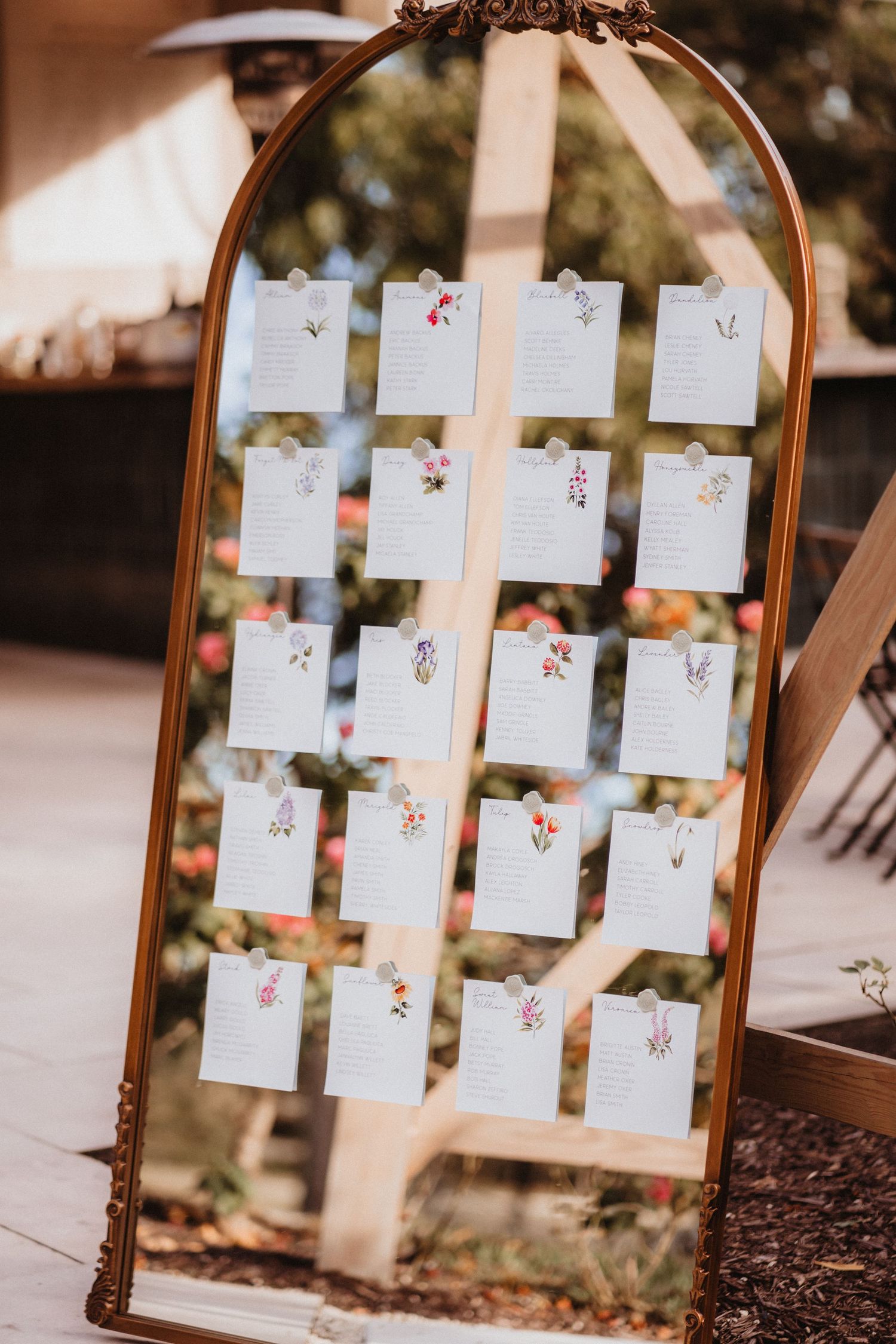 Ornate gold mirror displaying floral-illustrated seating chart cards for a wedding reception.