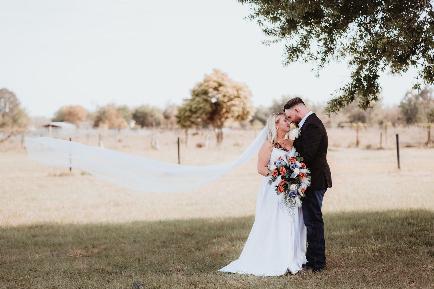 Bride and groom touching foreheads with veil flowing in wind holding colorful bouquet in open field.