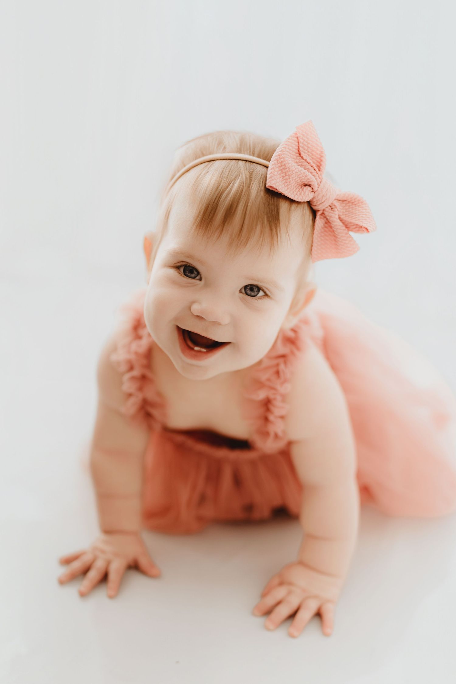 Happy smiling baby girl in pink tulle romper and bow headband crawling on bright white background.