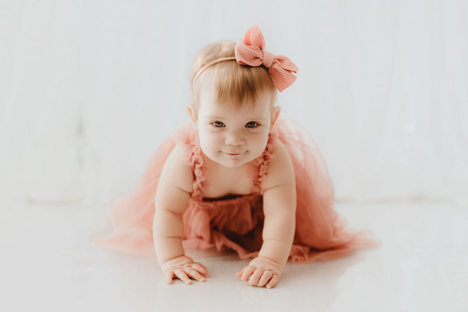 Cute baby girl in pink tulle dress and bow crawls on white floor.