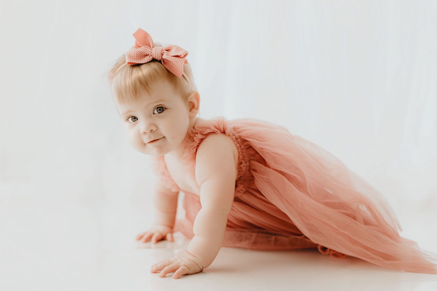 Adorable baby girl in pink tutu dress and bow headband crawling on white floor.