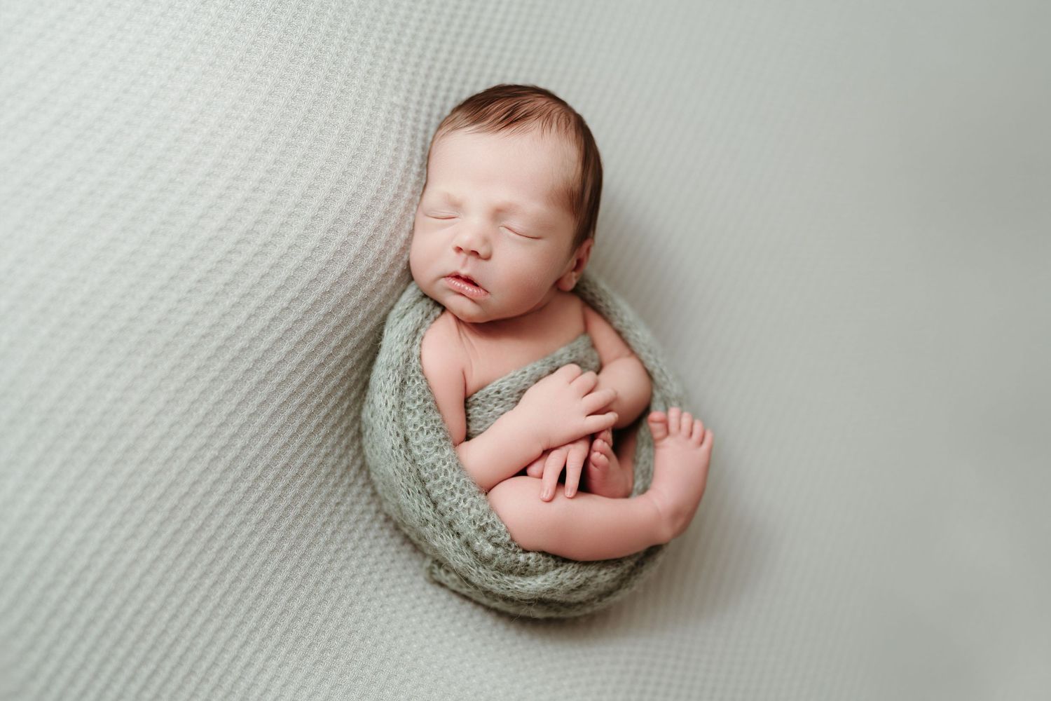 Newborn baby wrapped in green knit blanket sleeping in fetal position on gray surface.