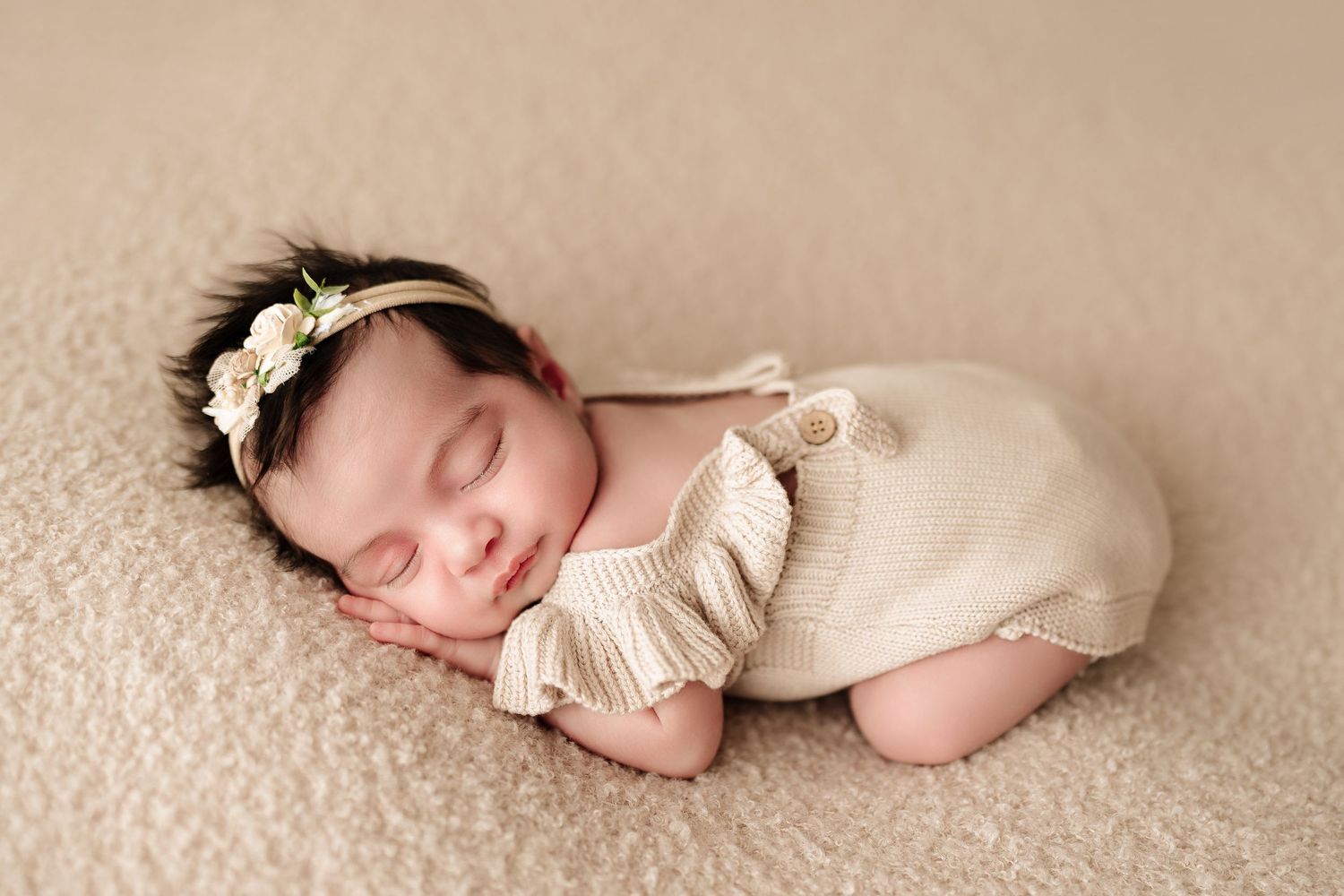 Peaceful sleeping newborn baby girl in beige knit romper and floral headband on soft blanket.